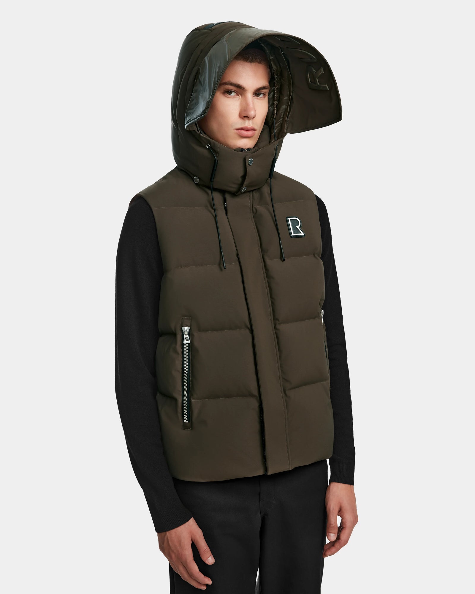 A man models the Rudsak REN M - 9123512 Army M puffer vest, weather-resistant with a high collar, large hood, R logo on the chest, and RDS-certified down insulation over a black shirt against a light gray background.