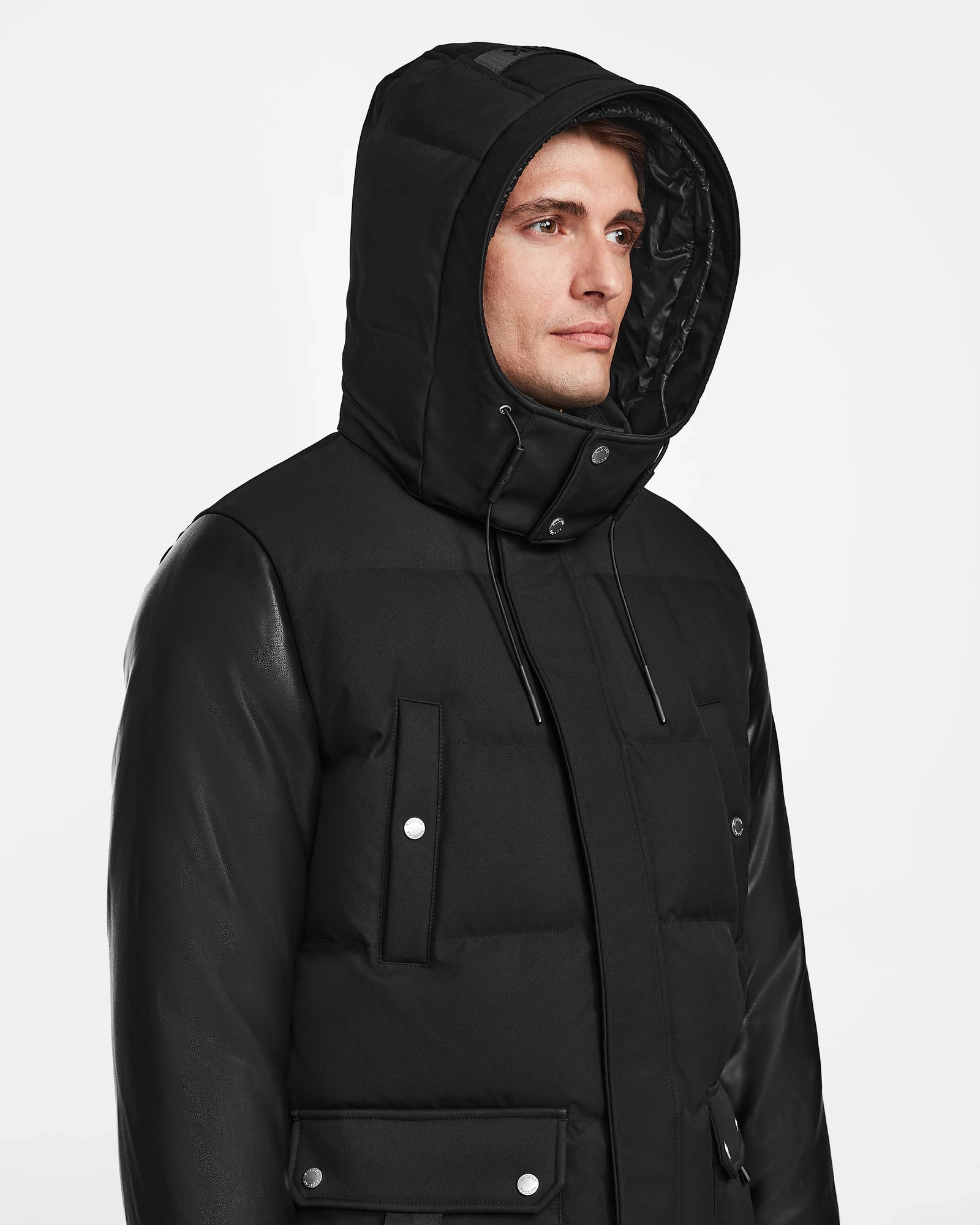 A man in the Rudsak RICKY R - M123506 black hooded puffer jacket with snap buttons and recycled leather sleeves stands against a plain white background, looking slightly to the side.