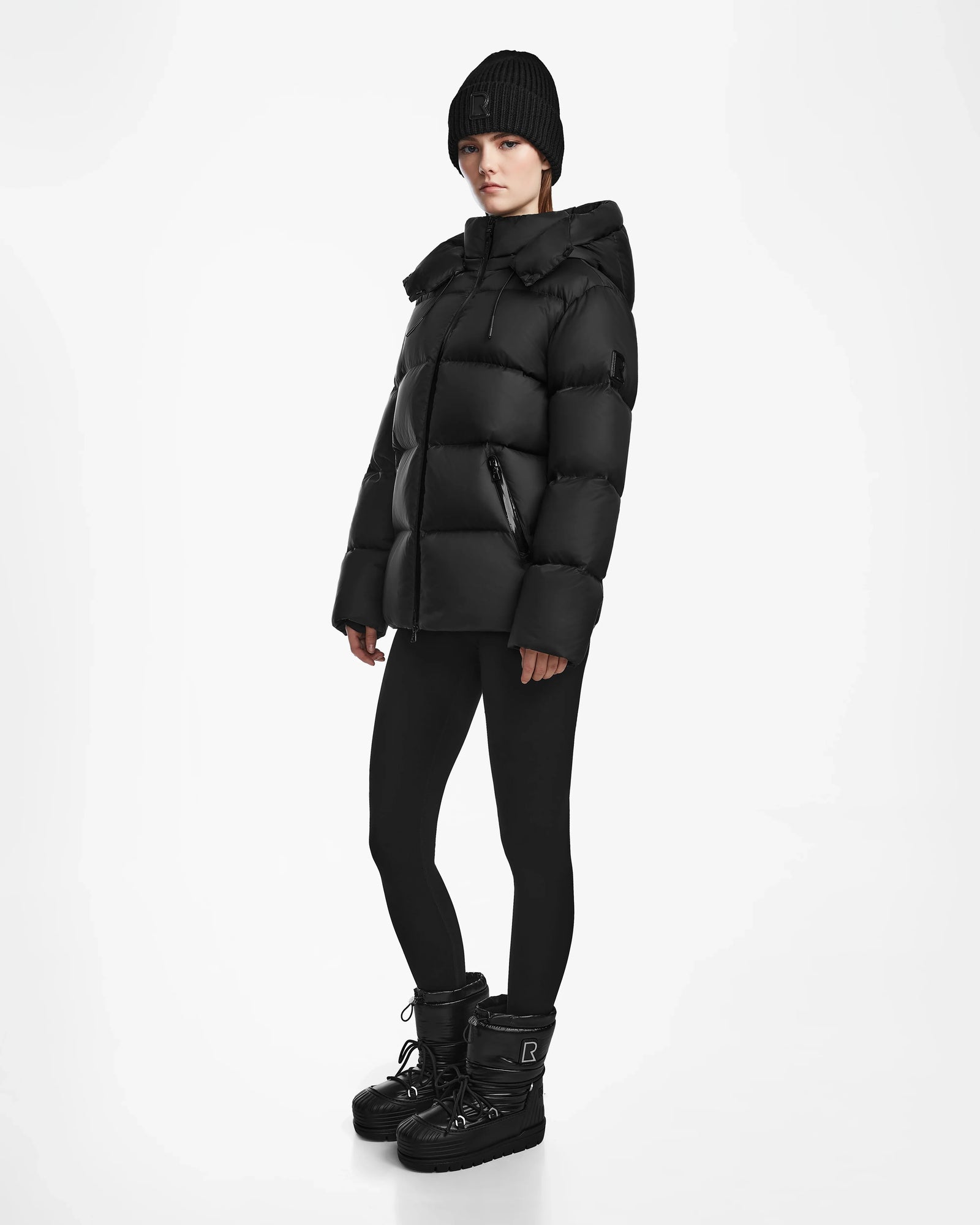 A person wearing the Rudsak RITA M - L124537 Black weather-resistant puffer jacket, black beanie, black leggings, and black winter boots stands against a plain white background.