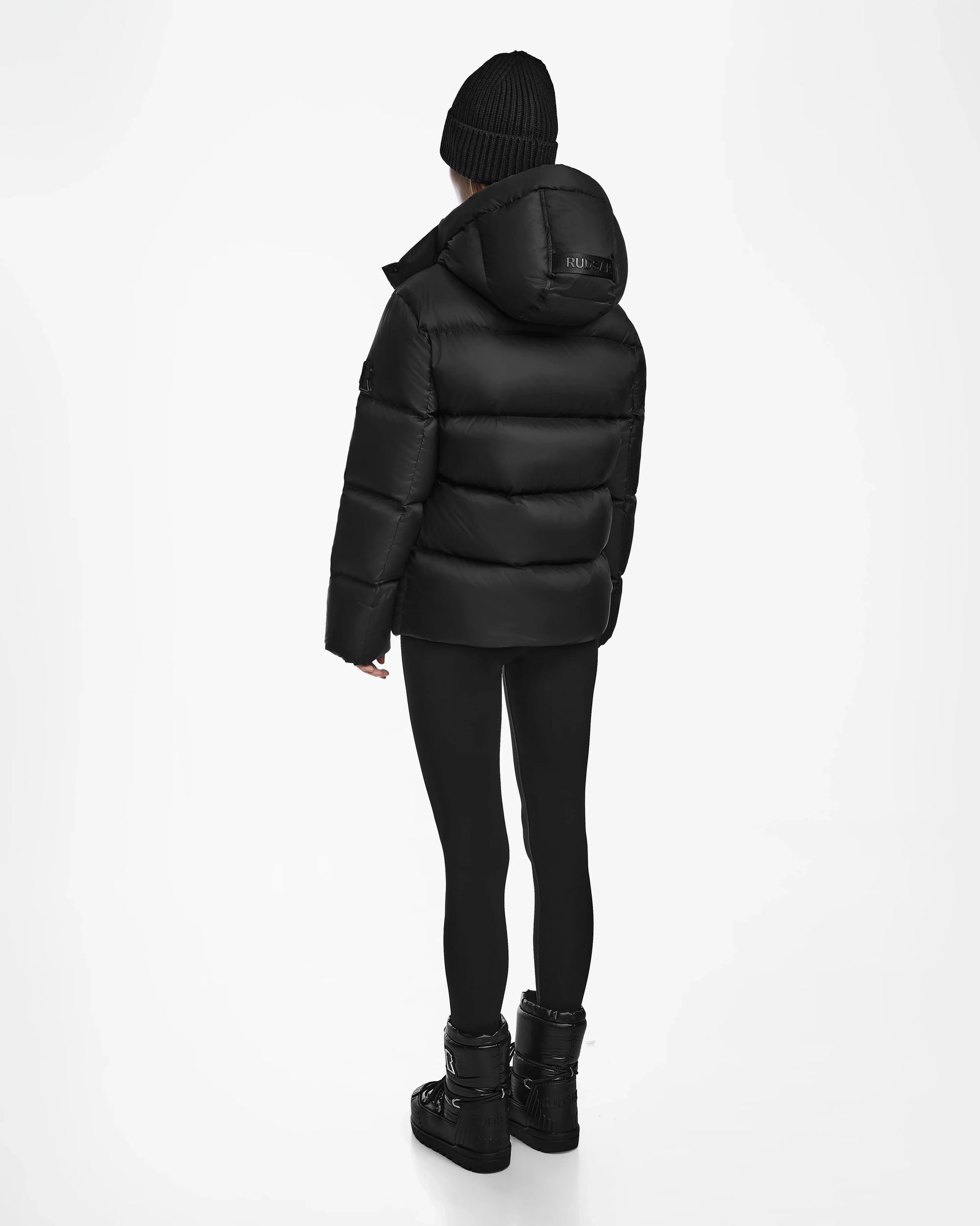 A person wearing the Rudsak RITA M - L124537 Black weather-resistant puffer, paired with a beanie, leggings, and boots, stands facing away on a white background.