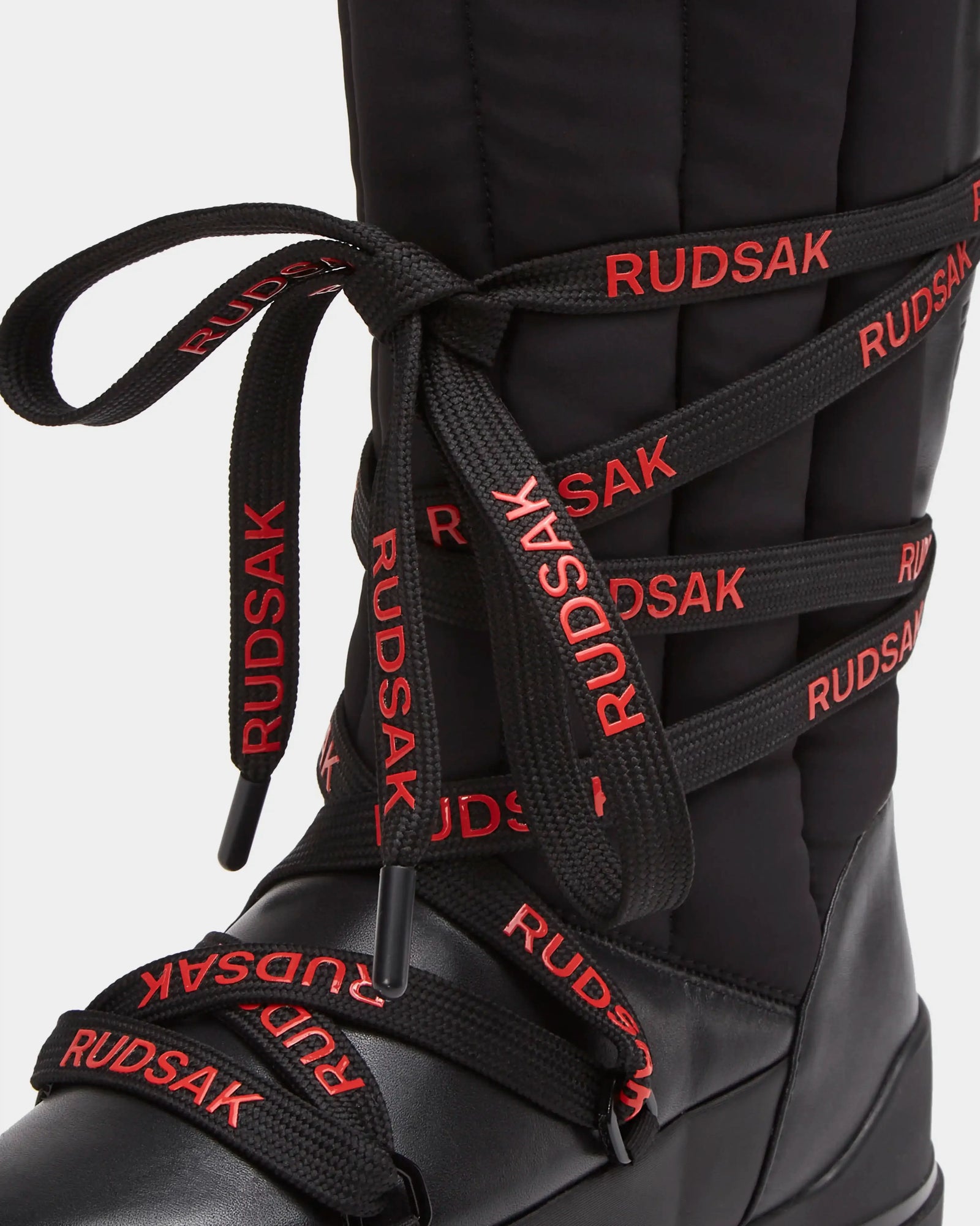 A close-up of the Rudsak RODIN - 8224502 Black womens winter boot shows thick black laces with repeating red RUDSAK text, a quilted texture, leather accents, and an anti-slip sole for secure steps.