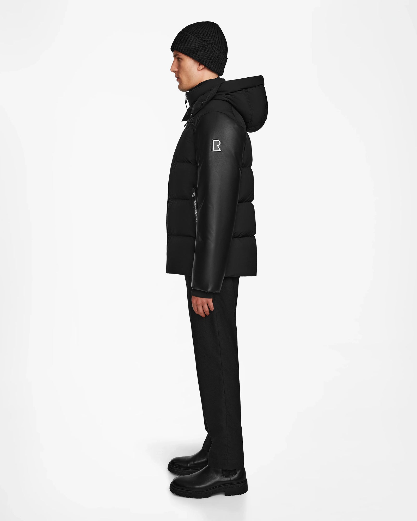 A person wearing the Rudsak RONNY M - M124513 Black puffer jacket stands sideways against a white background, paired with black pants, boots, and a knit beanie.