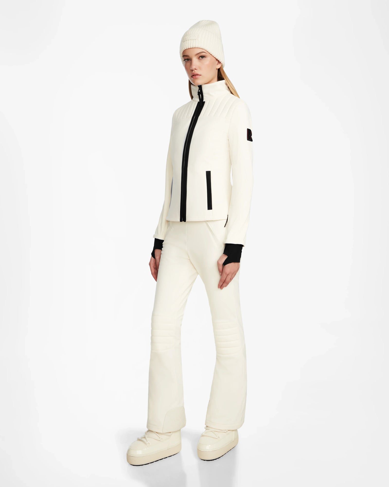A woman stands against a white background wearing the Rudsak ROSALIE - 8124784 Iceland women's fitted ski jacket with black zipper details, matching white pants, a white beanie, black gloves, and platform boots—perfect for stylish winter adventures.