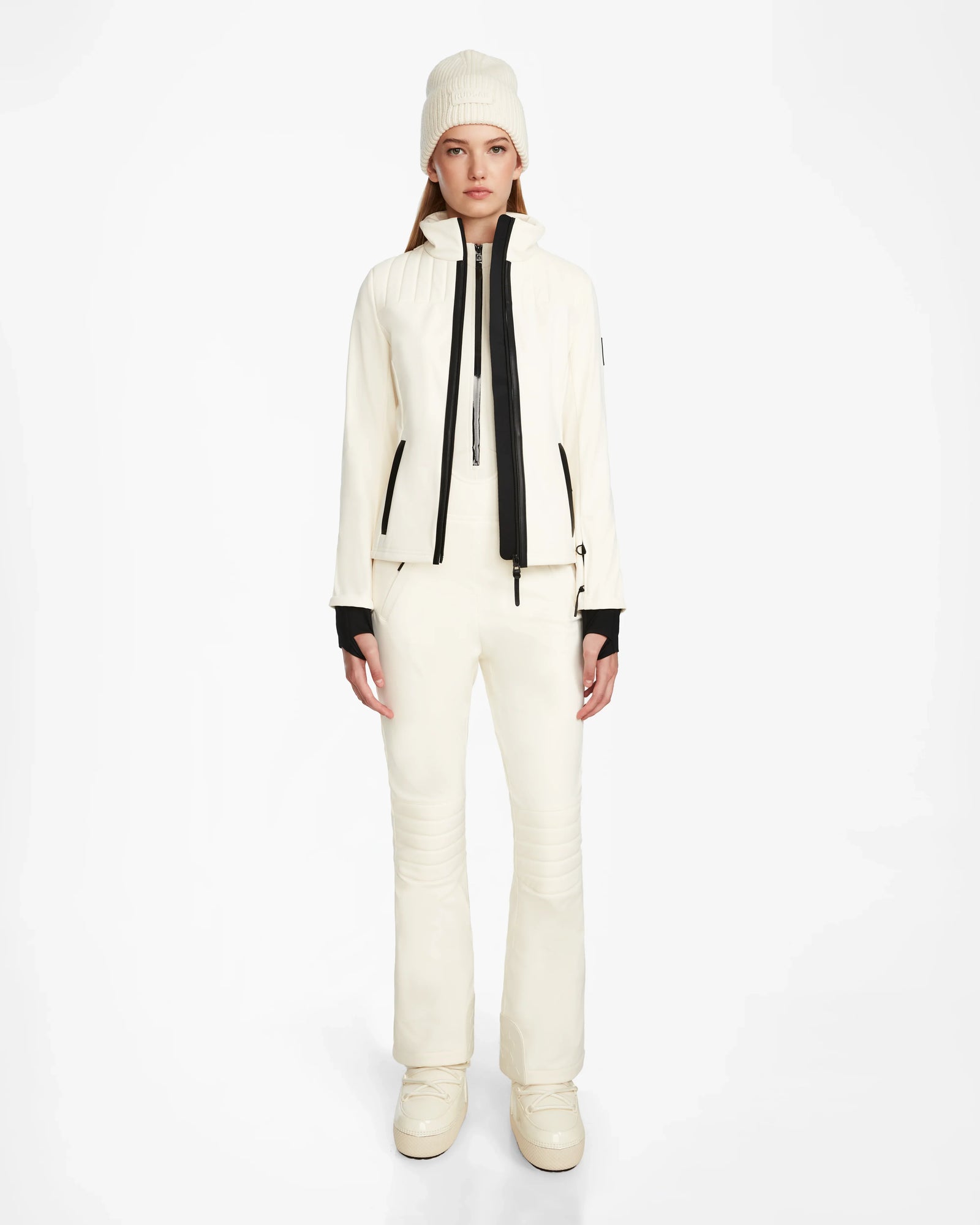 A woman faces forward in the Rudsak ROSALIE - 8124784 Iceland women’s ski jacket and matching pants, featuring black zipper and cuff accents. She completes the look with a knit beanie and boots against a plain white background.