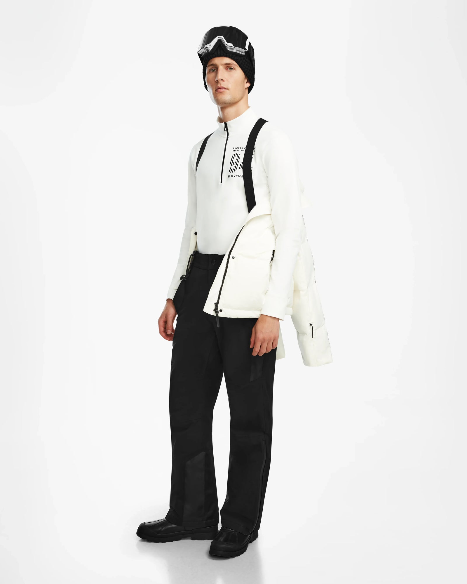 Dressed in black ski pants, boots, black goggles, a black hat, and a white long-sleeve top, the person wears Rudsaks USHER - 6124516 Optic White mens down ski jacket with RDS-certified insulation against a white background.