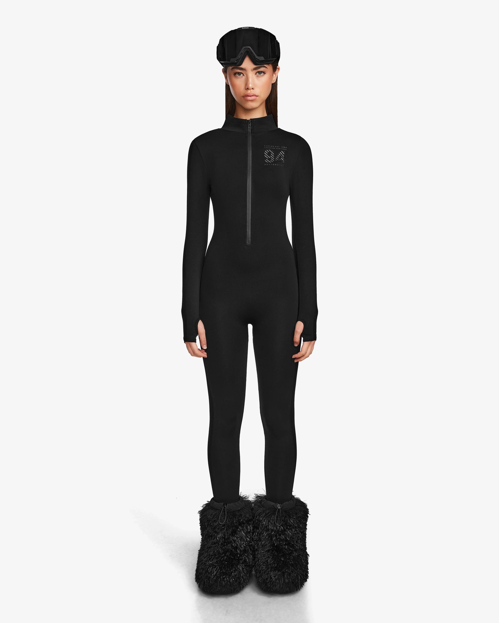 A woman faces forward in a fitted Rudsak ZADIE - 8524609 Black long-sleeve bodysuit with a front zipper, black fuzzy slippers, and a black sleep mask on her head, standing against a plain white background.