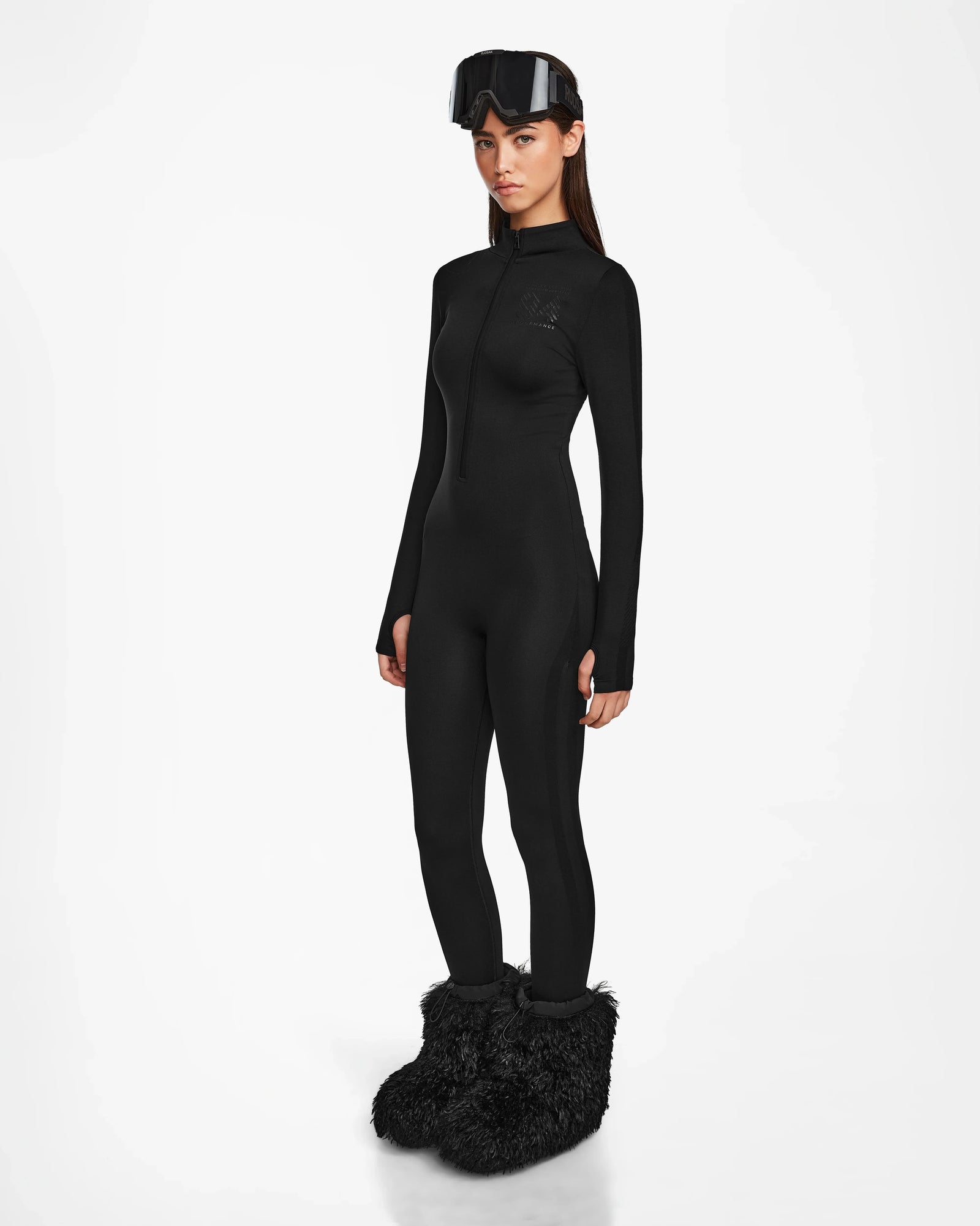 A woman poses against a white background in the Rudsak ZADIE - 8524609 Black ski catsuit, black ski goggles atop her head, and oversized black furry boots, with her arms resting by her sides.