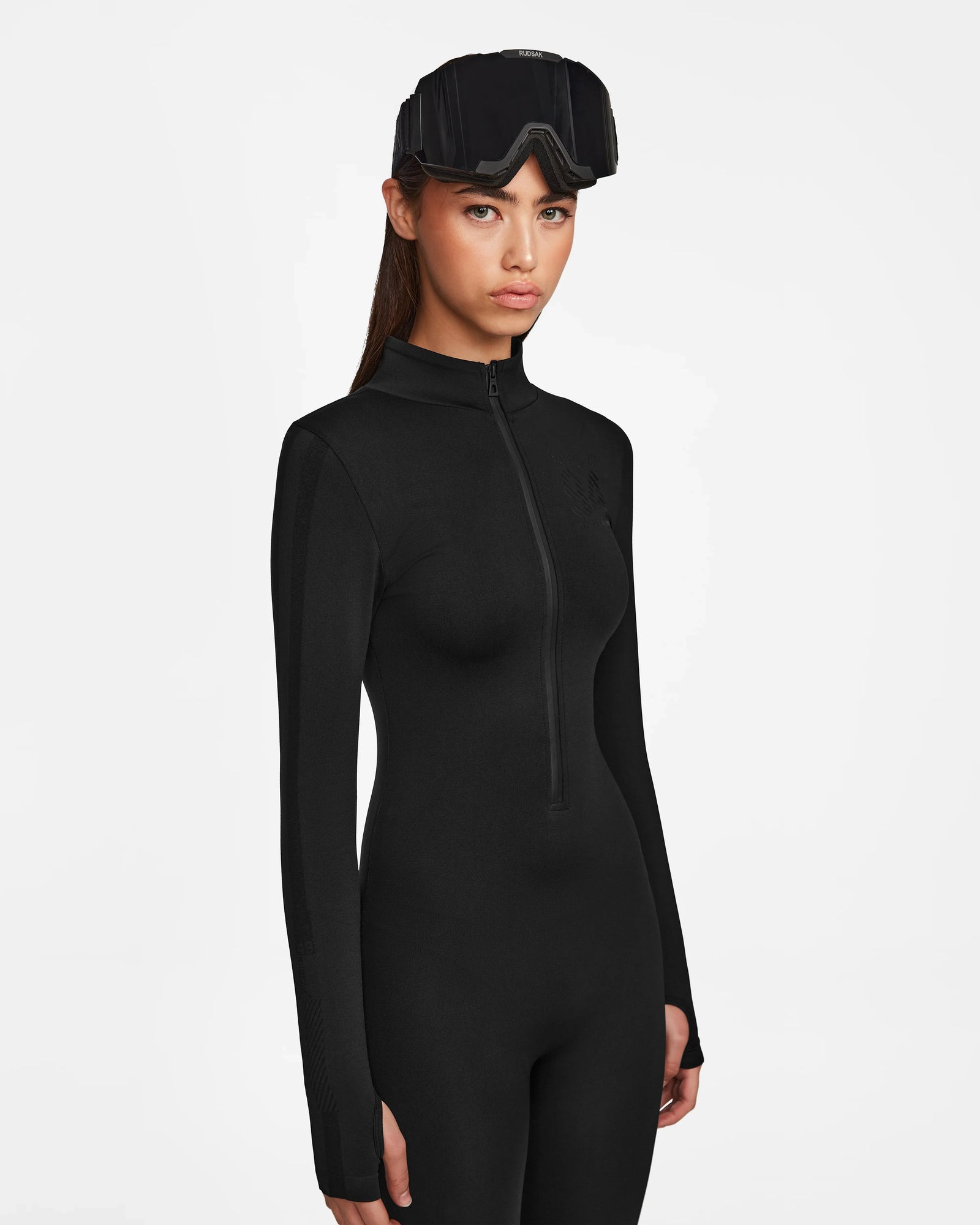 A woman in the ZADIE - 8524609 Black long sleeve bodysuit by Rudsak, wearing ski goggles on her head, stands against a plain white background with a neutral expression.