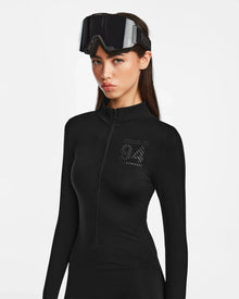 A woman stands against a plain white background wearing the black Rudsak ZADIE - 8524609 long-sleeve bodysuit with a subtle chest logo and large black ski goggles on her forehead.