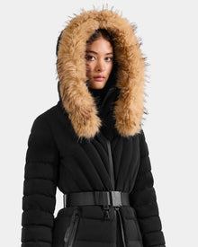 A woman models the Rudsak ALISA L - L125574 Black coat, a black quilted puffer with a belted waist and large hood trimmed with thick tan faux fur, posed against a plain light background.