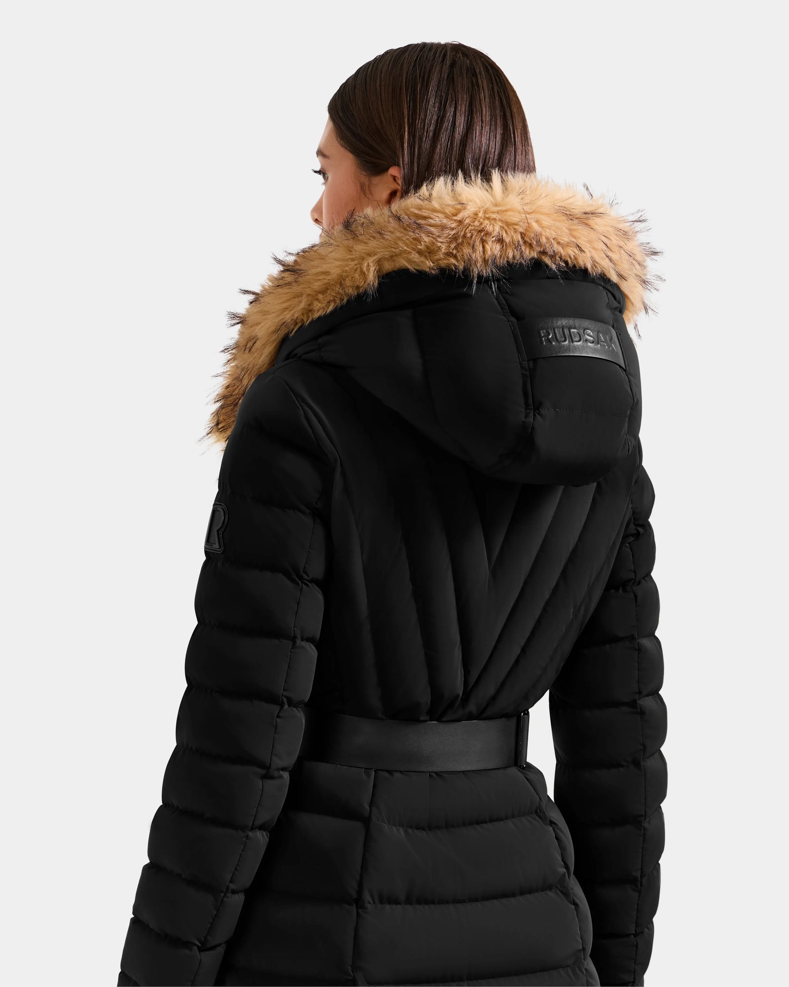 A person with straight brown hair is seen from behind wearing the Rudsak ALISA L - L125574 Black quilted puffer coat with a wide faux-fur trim hood and belt, set against a plain light background.