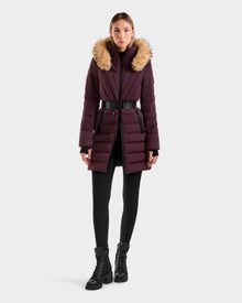 A woman faces forward, wearing the Rudsak ALISA M - L125586 Bordeaux quilted down parka with a vegan leather belt and faux fur hood, paired with black leggings and black lace-up boots against a plain white background.
