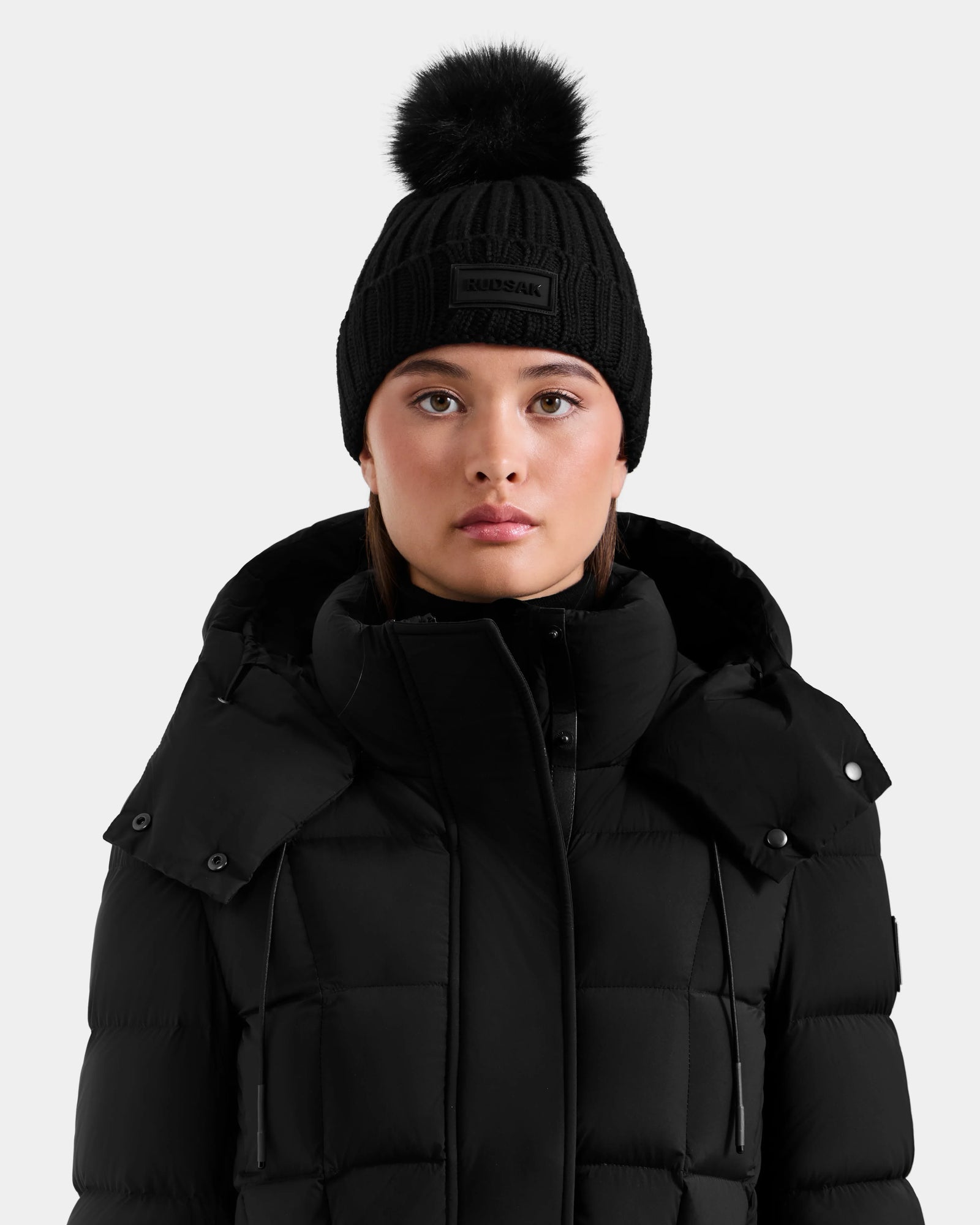 A woman wears the Rudsak ANISA - L423500 Black/Black puffer jacket and a matching black knit pom pom hat, standing against a plain light background and looking at the camera—an ideal addition to any winter wardrobe.