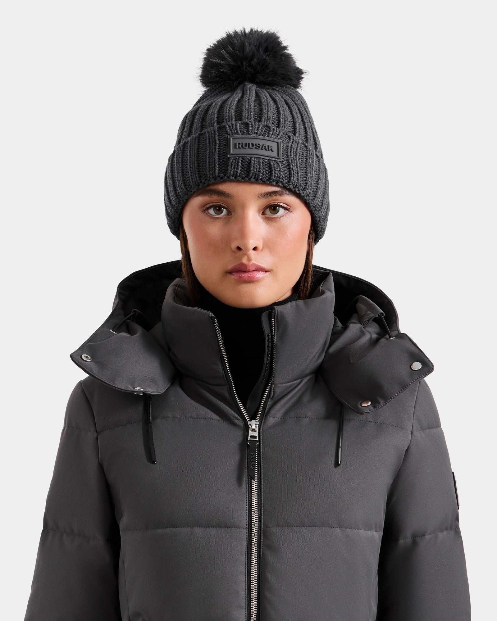 A person wears the Rudsak ANISA - L423500 Grey dark gray hooded puffer jacket and a women’s knit hat with a black faux fur pom pom, facing forward against a plain light background—ideal for winter style.