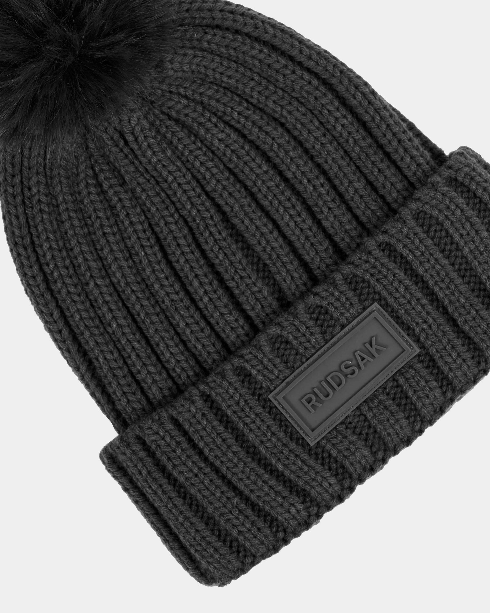 A close-up of the Rudsak ANISA - L423500 Grey ribbed knit beanie with a fold-over cuff, large faux fur pom pom, and signature RUDSAK patch—an essential winter accessory.