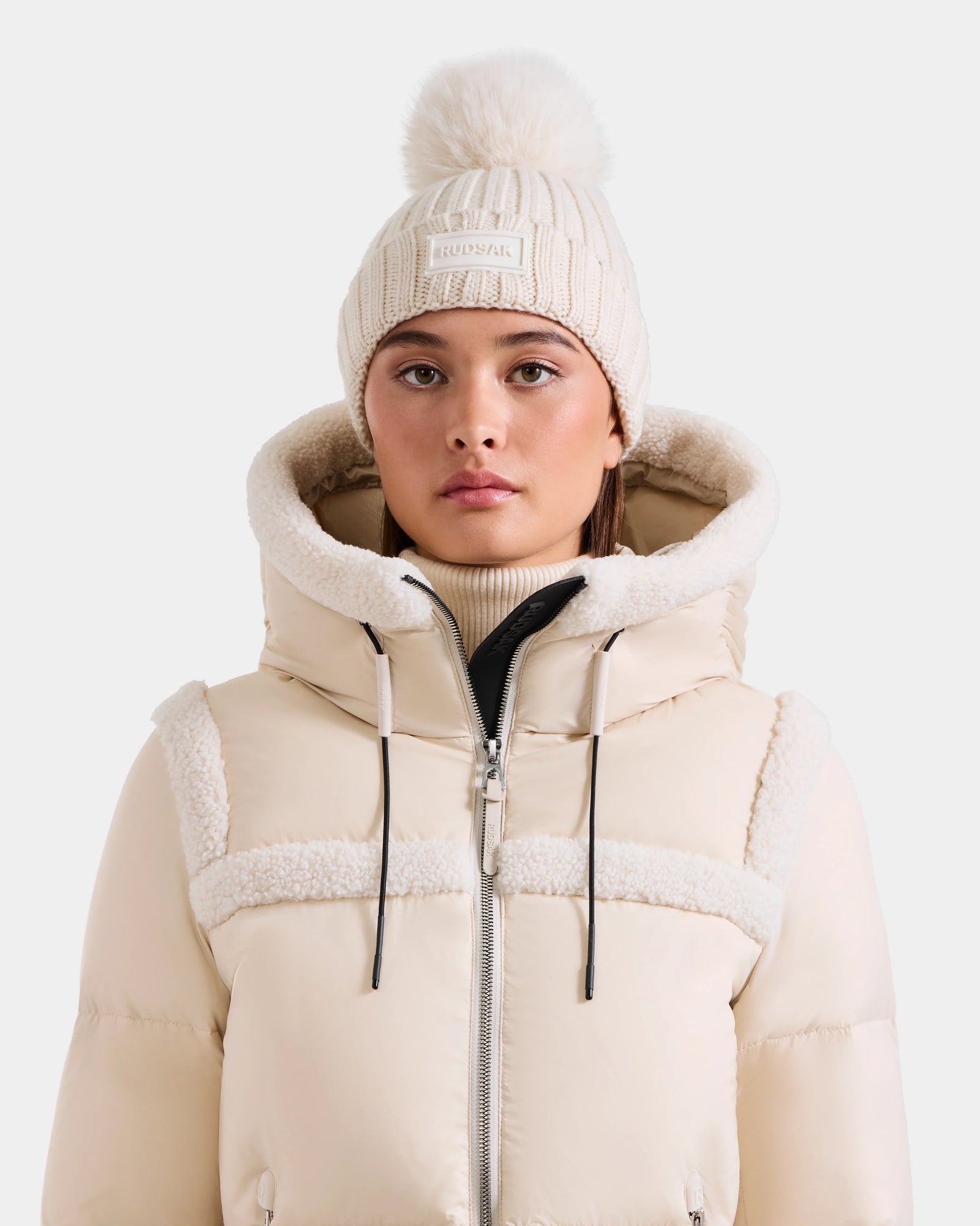 A woman wears the Rudsak ANISA - L423500 Vanilla beige puffer jacket with sherpa-lined hood and a cream knit pom pom hat, standing against a plain background—perfect cozy pieces for any winter wardrobe.