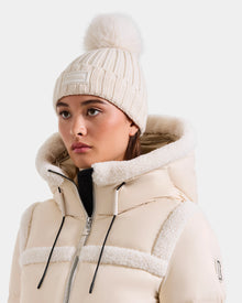 A woman showcases cozy winter style in the Rudsak ANISA - L423500 Vanilla, a cream knit pom pom hat with faux fur detail, paired with a matching high-collared puffer jacket, glancing to the side against a plain background.