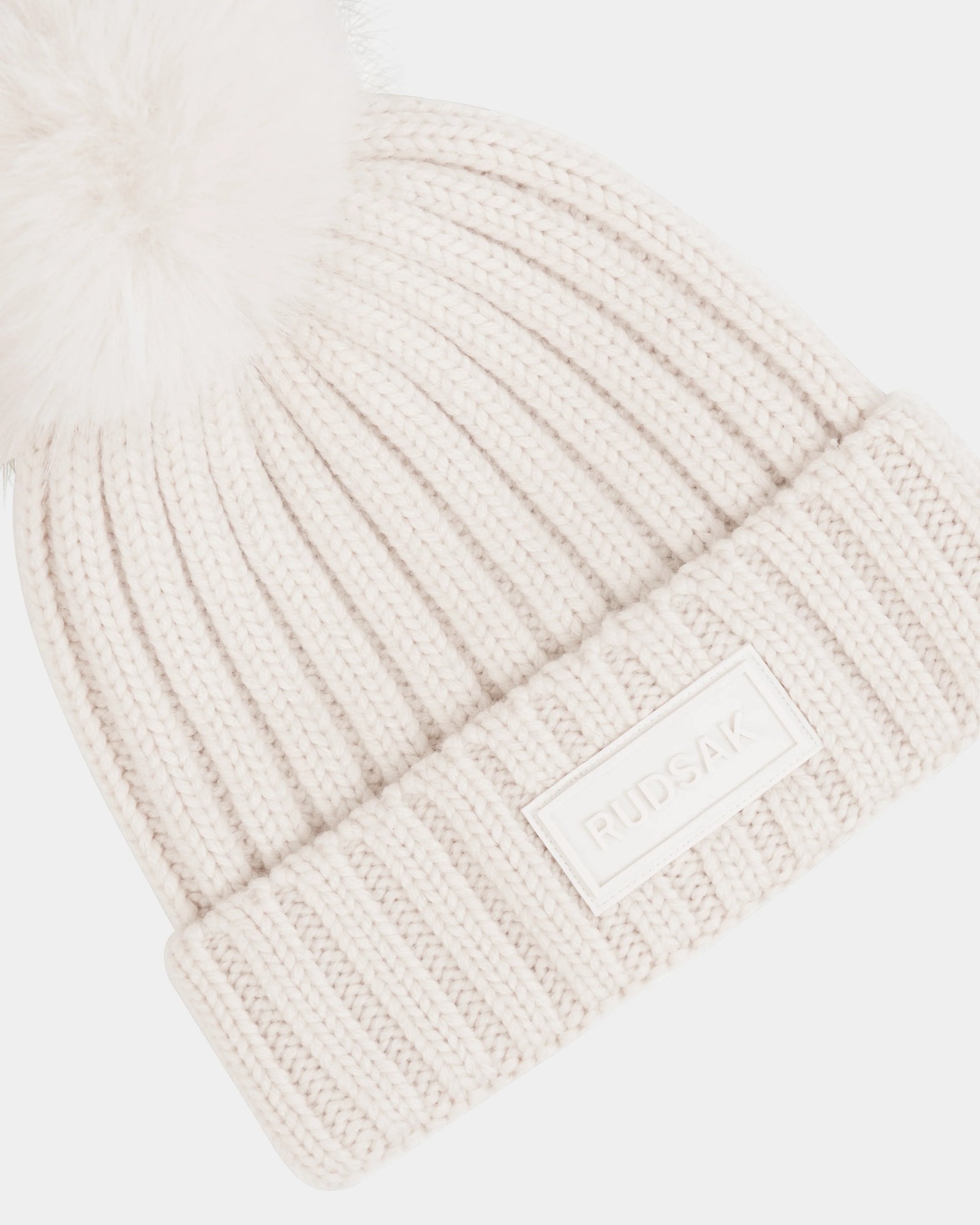 The ANISA - L423500 Vanilla by Rudsak is a cream ribbed knit beanie featuring a large faux fur pom pom and a folded brim with a rectangular Rudsak logo patch—an essential addition to your winter wardrobe.