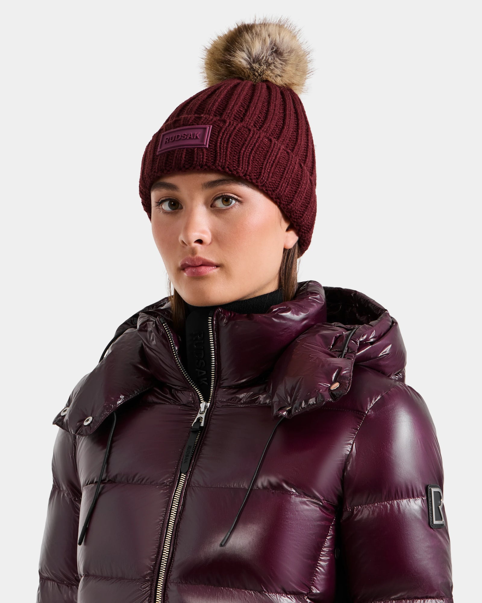Wearing the Rudsak ANISA - L423500 Wine women’s knit pom pom hat with faux fur and a shiny, dark purple puffer jacket, she looks forward with a neutral expression—an ideal pick for your winter wardrobe.