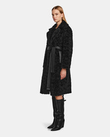 A woman in profile wears the Rudsak ANNABELLE - L124544 Black vegan leather coat with a black belt, black knee-high boots, and straight blonde hair, set against a plain white background.