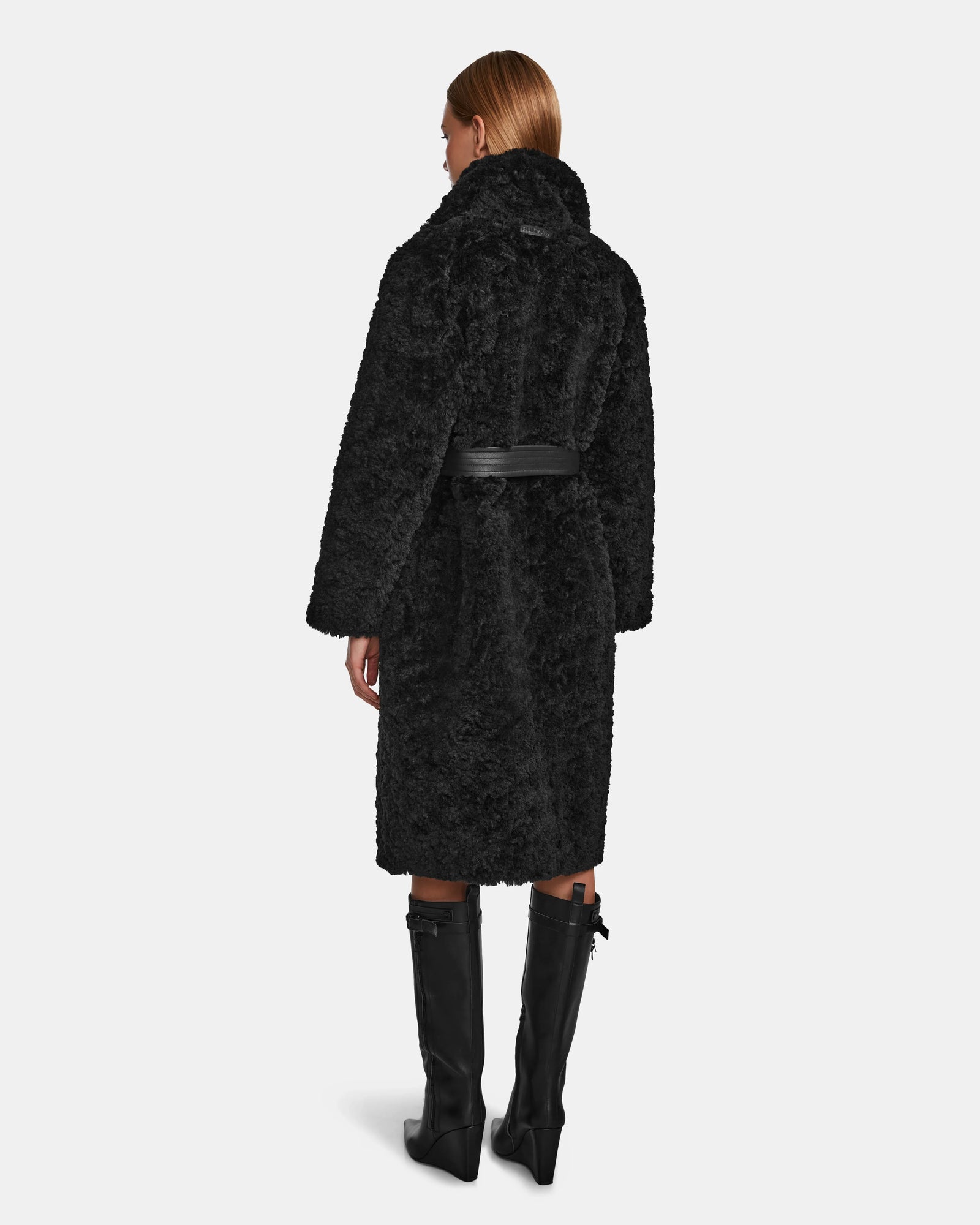 A woman with straight, light brown hair is seen from the back wearing the Rudsak ANNABELLE - L124544 Black faux fur coat with a black belt and tall black boots against a plain white background.
