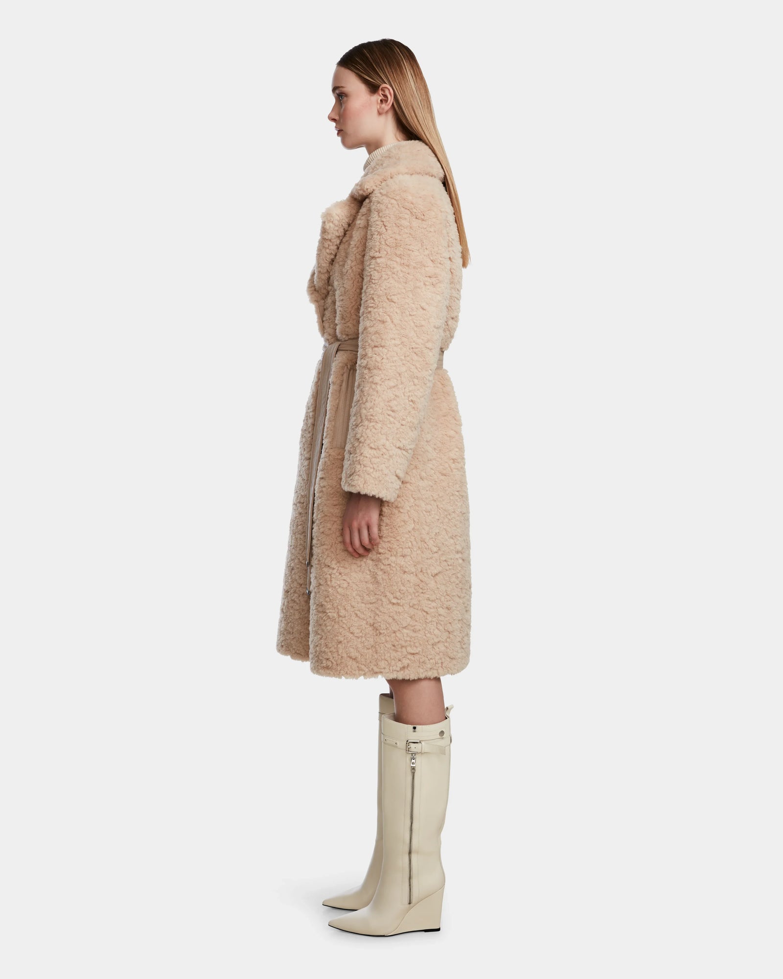 A woman stands in profile against a white background, wearing the Rudsak ANNABELLE - L124544 Sand knee-length reversible teddy coat and tall, off-white heeled boots with side zippers.