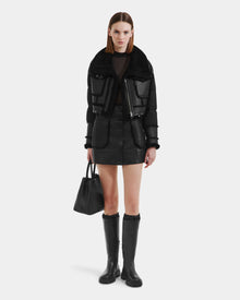 A woman with straight, shoulder-length hair and a neutral expression models the Rudsak ARIELLA - 8125505 Black shearling jacket, styled with a black mini skirt, knee-high boots, and a handbag against a plain background.