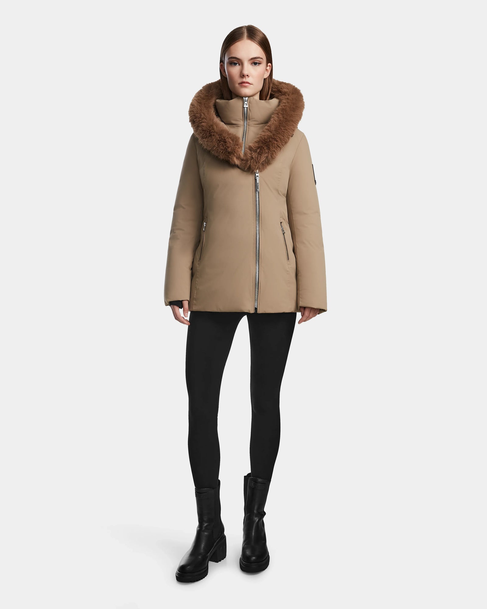 A woman stands facing forward, wearing the Rudsak AURORE - L123561 Sand fitted winter coat with a large fur-trimmed collar, paired with black leggings and black boots against a plain white background.