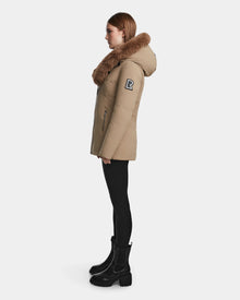 A woman stands in profile against a plain white background, wearing the Rudsak AURORE - L123561 Sand winter parka with a large fur-lined hood, black leggings, and boots. The fitted coat features an 