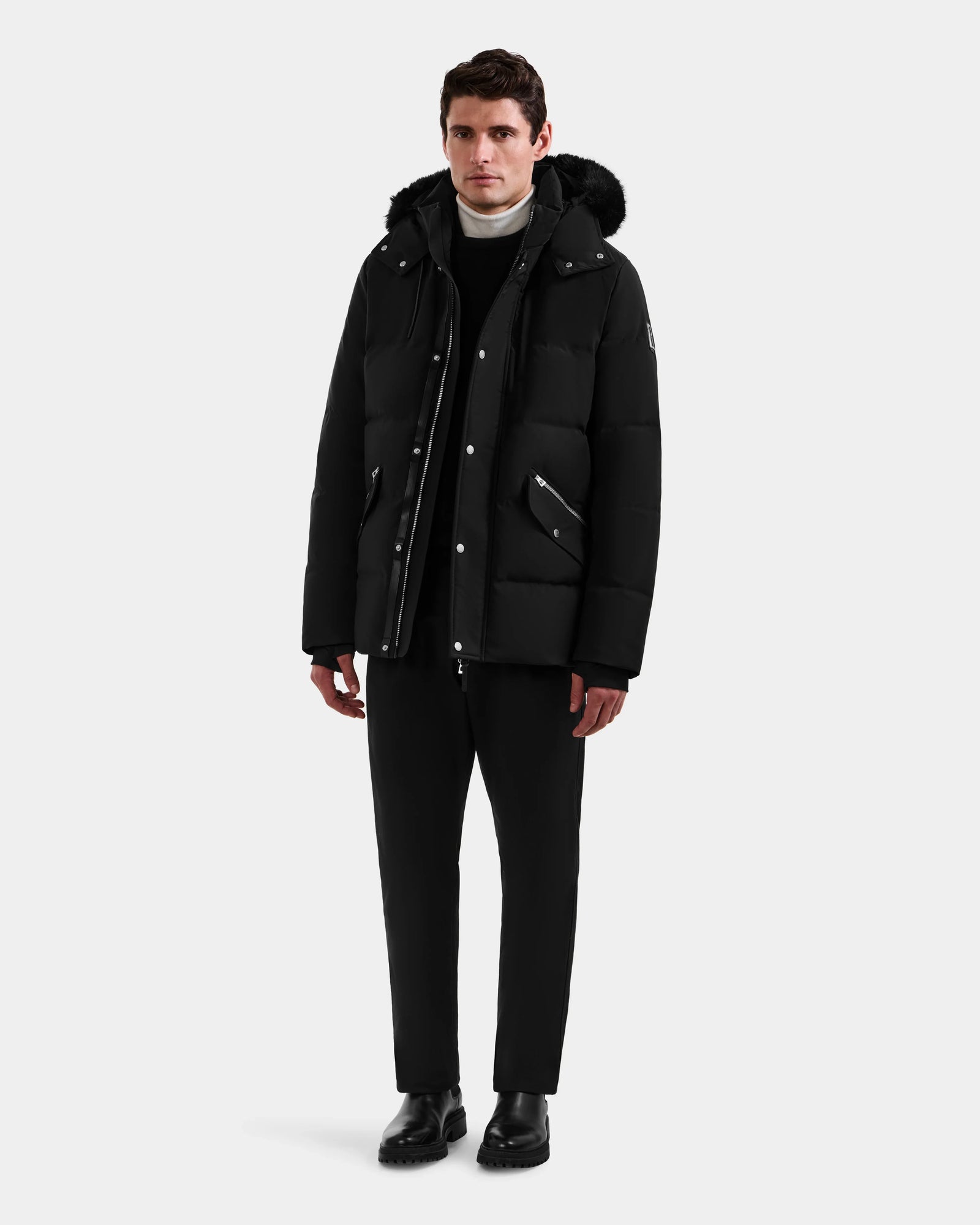A man faces forward wearing the Rudsak BRADY - M125510 Black men’s quilted down parka with removable hood, paired with black pants, black shoes, and a white turtleneck against a plain light gray background.