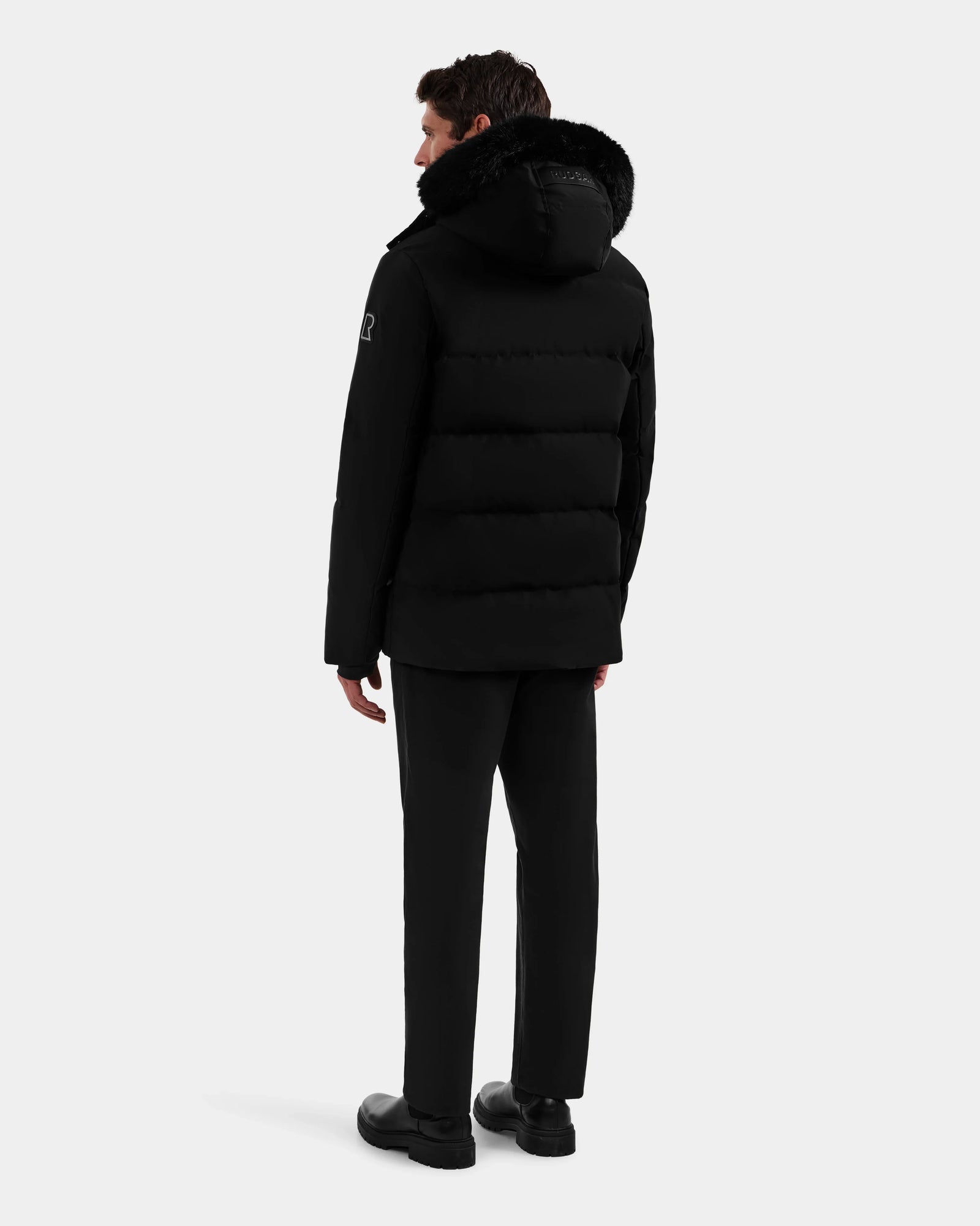 A person faces away, wearing the Rudsak BRADY - M125510 Black men’s quilted down parka with a fur-trimmed removable hood, paired with black pants and shoes, set against a plain white background—a true winter essential.