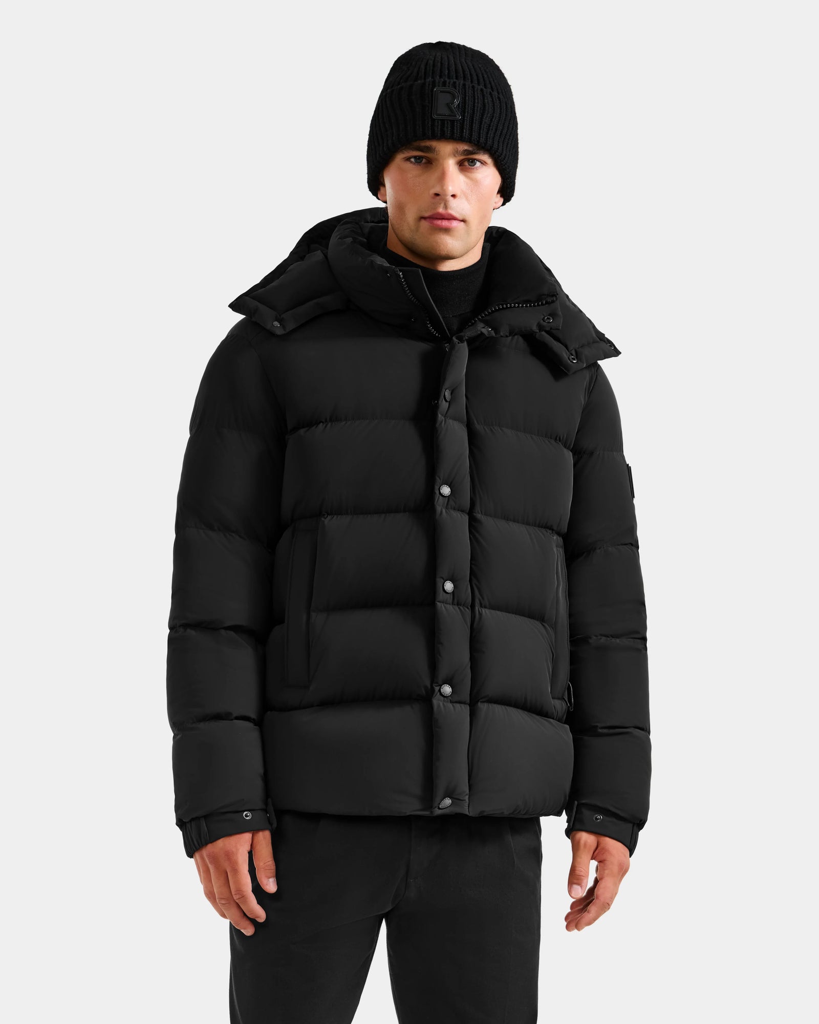 A person wearing a black beanie and the Rudsak BRAYDEN M-S - M125524 Black men’s puffer jacket with a hood stands facing forward against a plain light gray background.