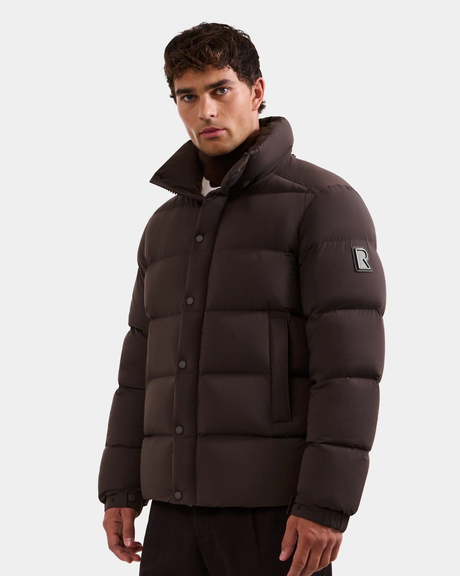 A man wearing the Rudsak BRAYDEN M-S - M125524 Espresso down puffer jacket with a high collar and an R patch on the upper sleeve stands against a plain, light background.