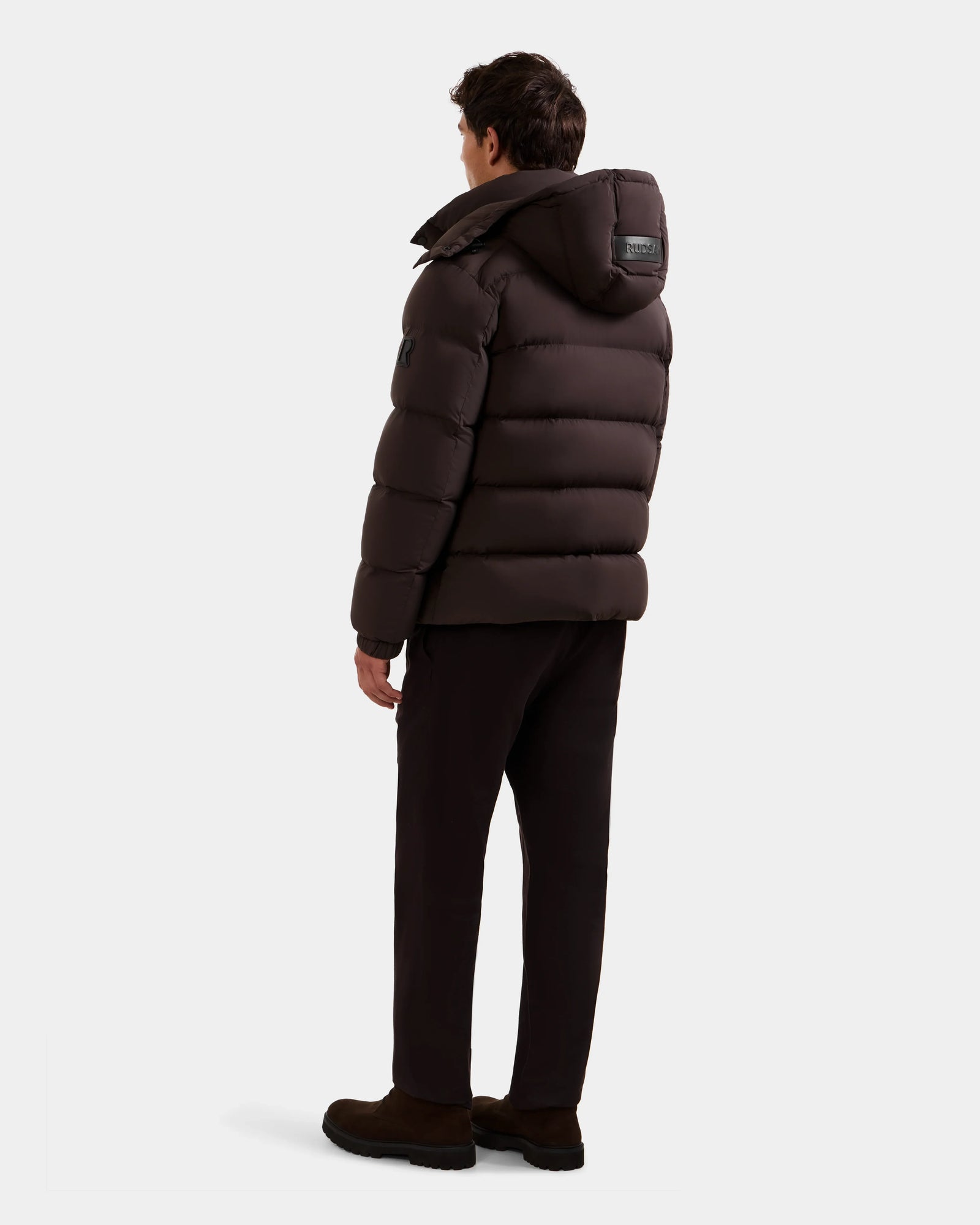 A man stands facing away, wearing the Rudsak BRAYDEN M-S - M125524 Espresso RDS-certified down winter jacket with a hood, paired with dark pants and shoes, set against a plain light background.