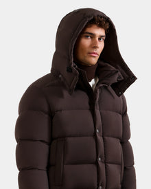 A man wearing the Rudsak BRAYDEN M-S – M125524 Espresso down puffer with a hood, buttoned up, poses warmly dressed against a plain light background.