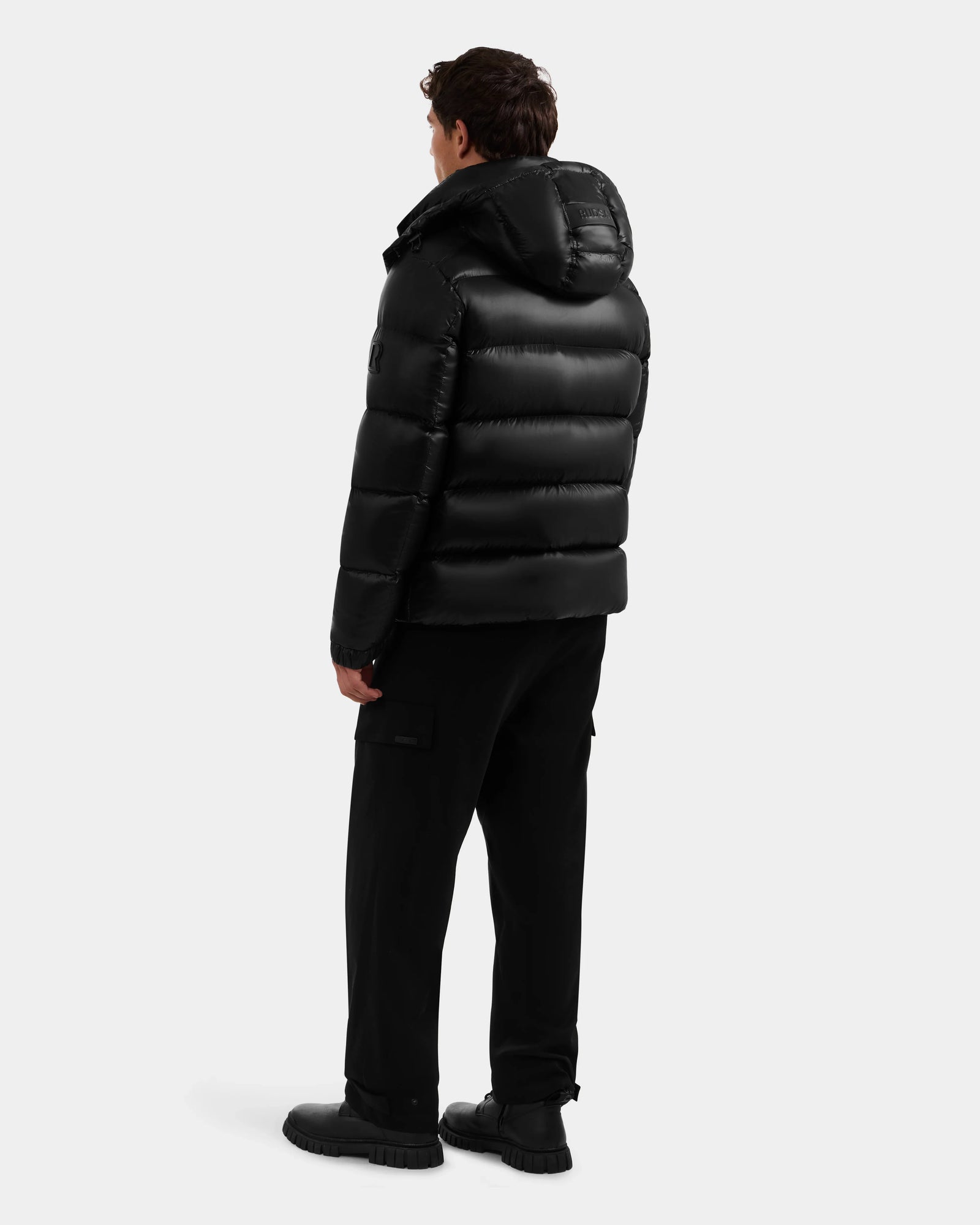 A person faces away, wearing the Rudsak BRAYDEN S - M125525 Black men's quilted down puffer jacket with hood, paired with black pants and shoes against a plain light background.