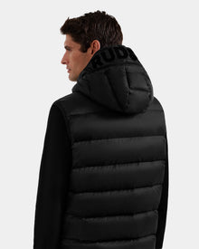 A man with short brown hair is seen from the back wearing the Rudsak BROOKS - M125520 Black puffer jacket, featuring a hood crafted from recycled-nylon shell with bold black lettering. The background is plain and light-colored.