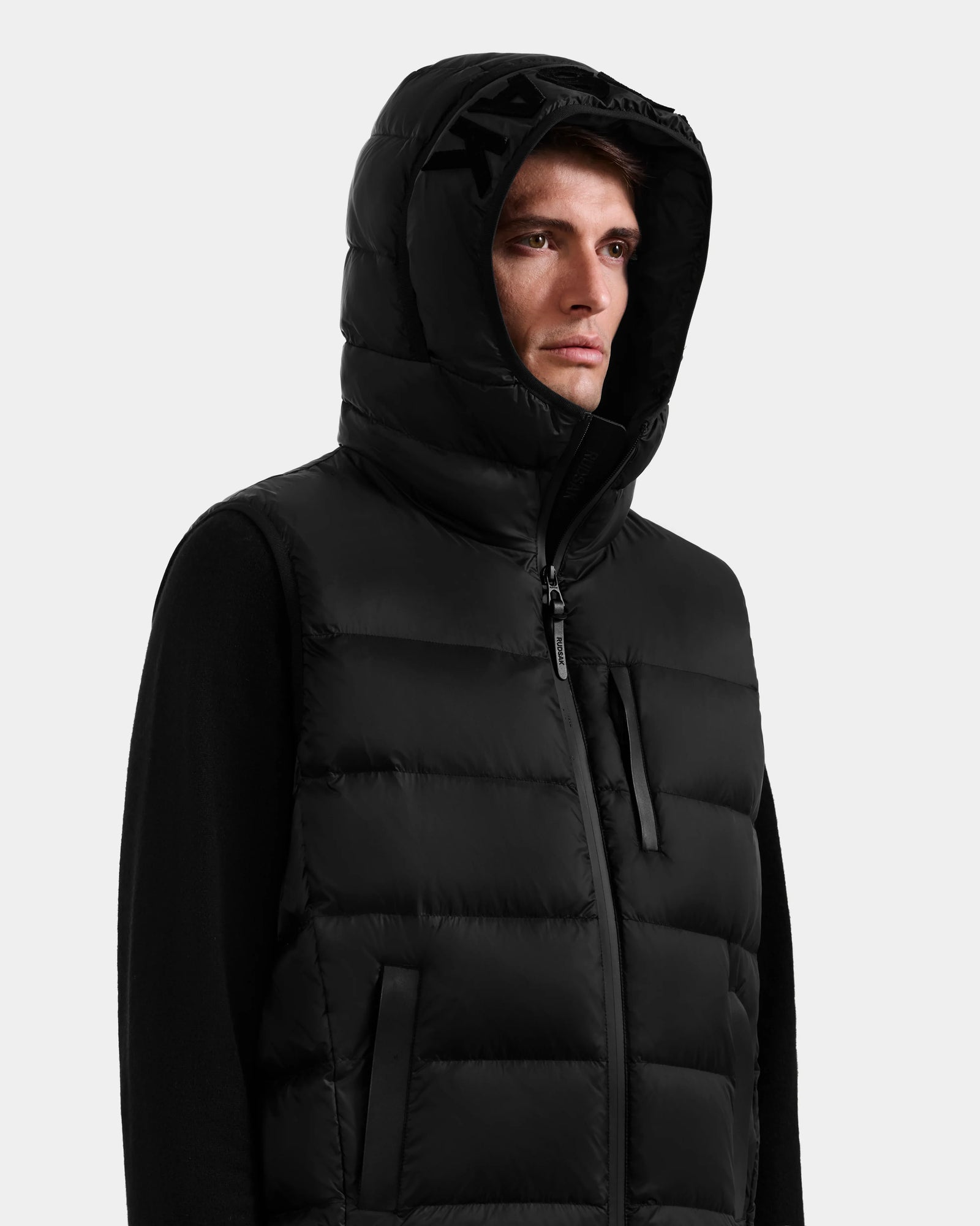 A person wears the Rudsak BROOKS - M125520 Black hooded down vest made from recycled-nylon shell, zipped up with the hood on, looking to the side against a plain light background.