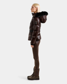 A woman with straight blonde hair stands in profile wearing the Rudsak CHRISTELLE S - L125544 Espresso women's shiny dark brown down puffer jacket with a faux fur trim hood, against a plain light background.