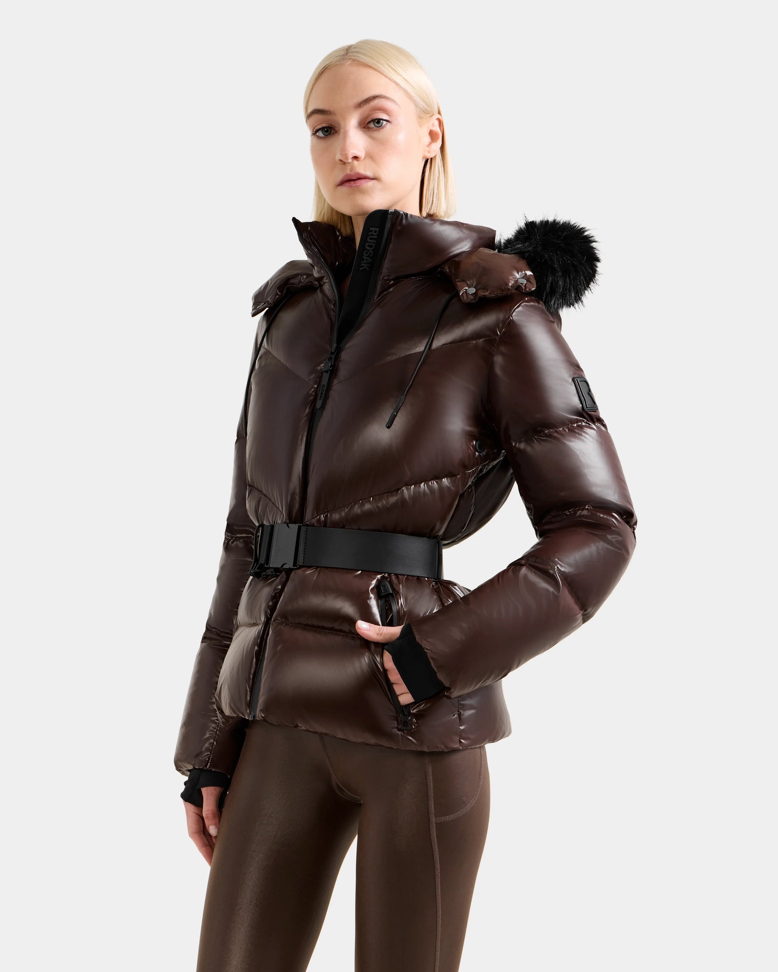 A woman with straight blonde hair models the Rudsak CHRISTELLE S - L125544 Espresso, a glossy women’s down puffer jacket with hood, black faux fur trim, and belted waist, paired with brown leggings against a light gray background.