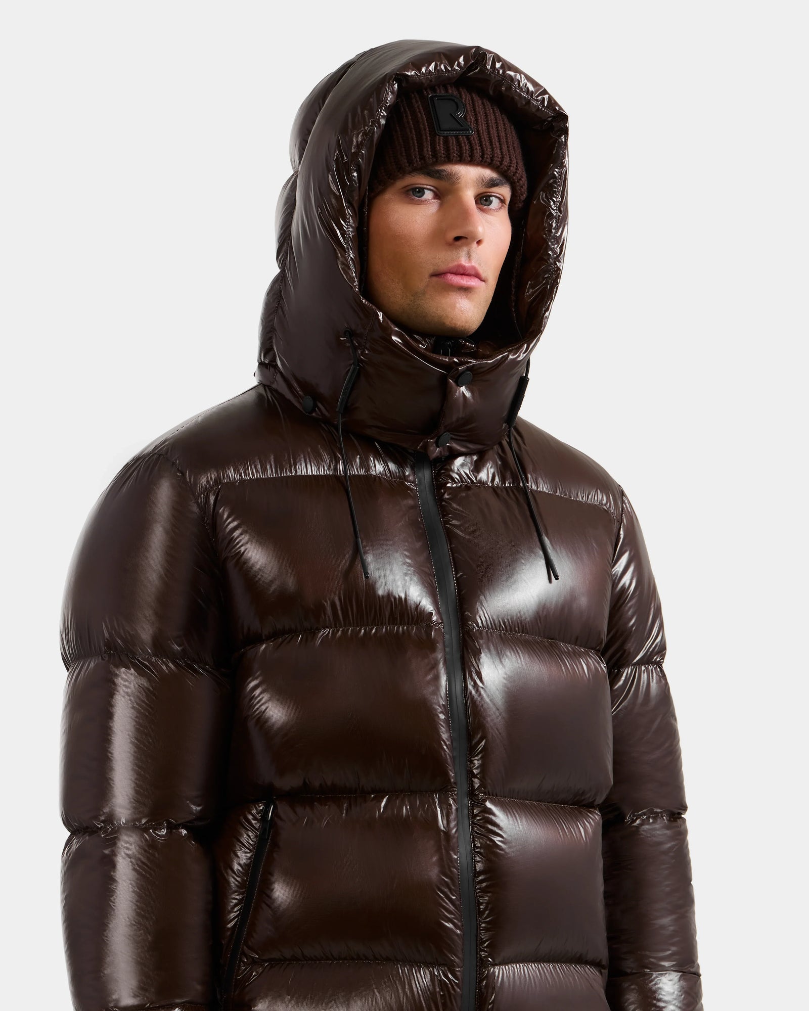 A person wears the Rudsak DANIEL S - M124508 Espresso men’s hooded down puffer with a glossy, weather-resistant shell, high-loft down fill, high collar, black zipper, hood up, and a dark knit beanie against a plain white background.