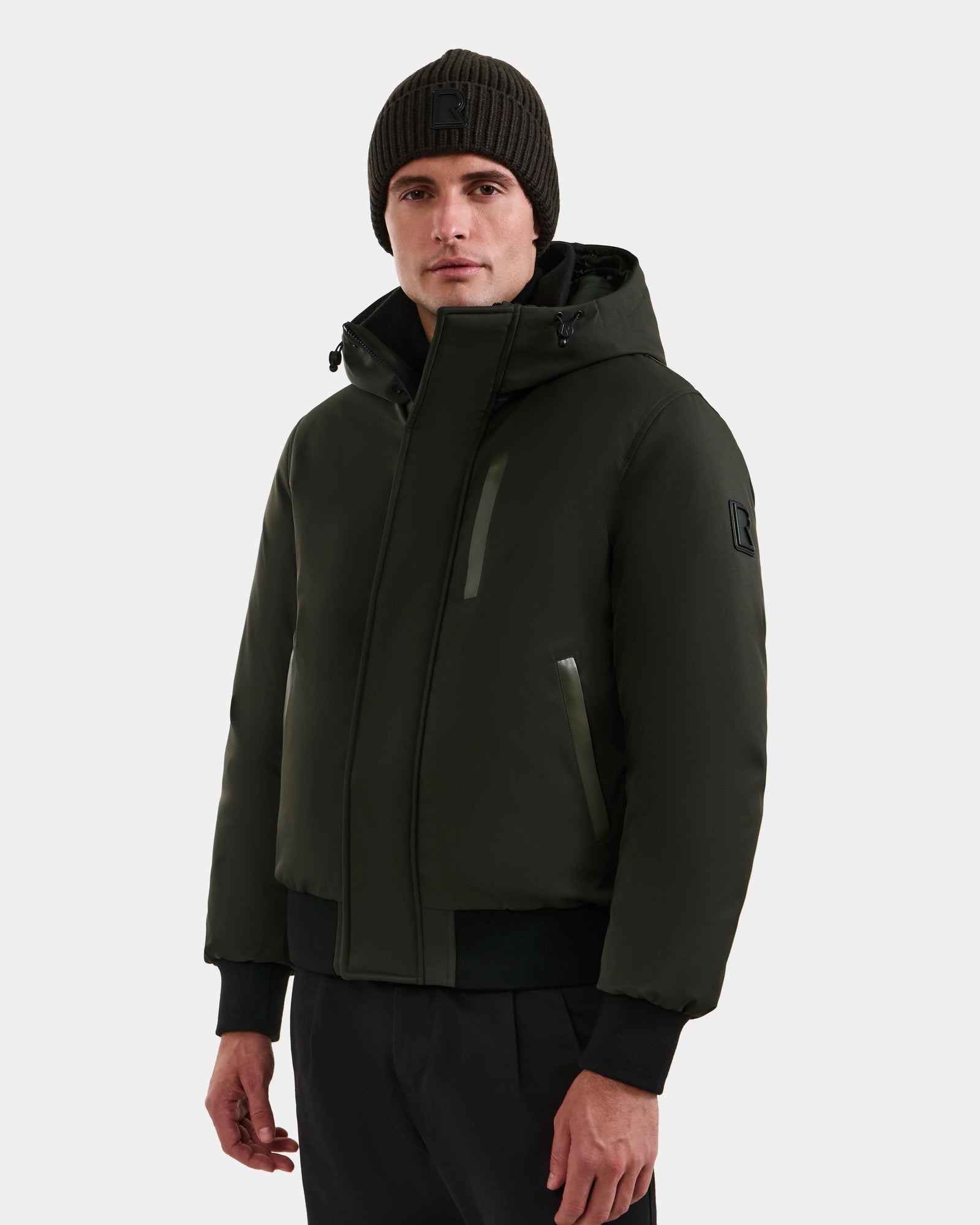 A man wears the Rudsak DEACON - M123513 Dark Olive men’s down puffer jacket with a hood, paired with a black knit beanie and black pants, standing against a plain light gray background.