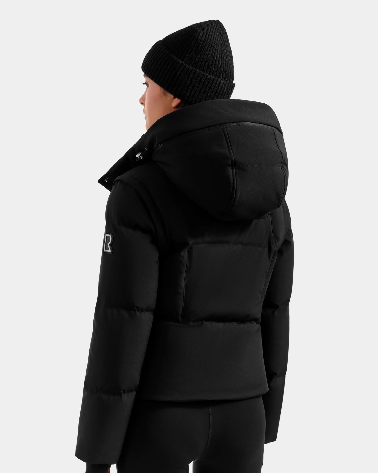 A person wearing a black Rudsak DELILAH M down puffer with hood and a black beanie, seen from the back against a light background. The jacket has an "R" patch on the upper sleeve.