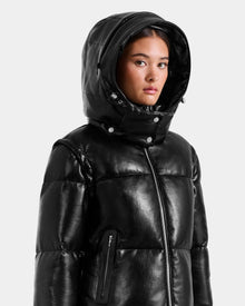 A young woman wears the Rudsak DELILAH PX - L125537 black vegan-leather puffer jacket with a high zip-up collar and hood, looking slightly to the side against a plain, light background.