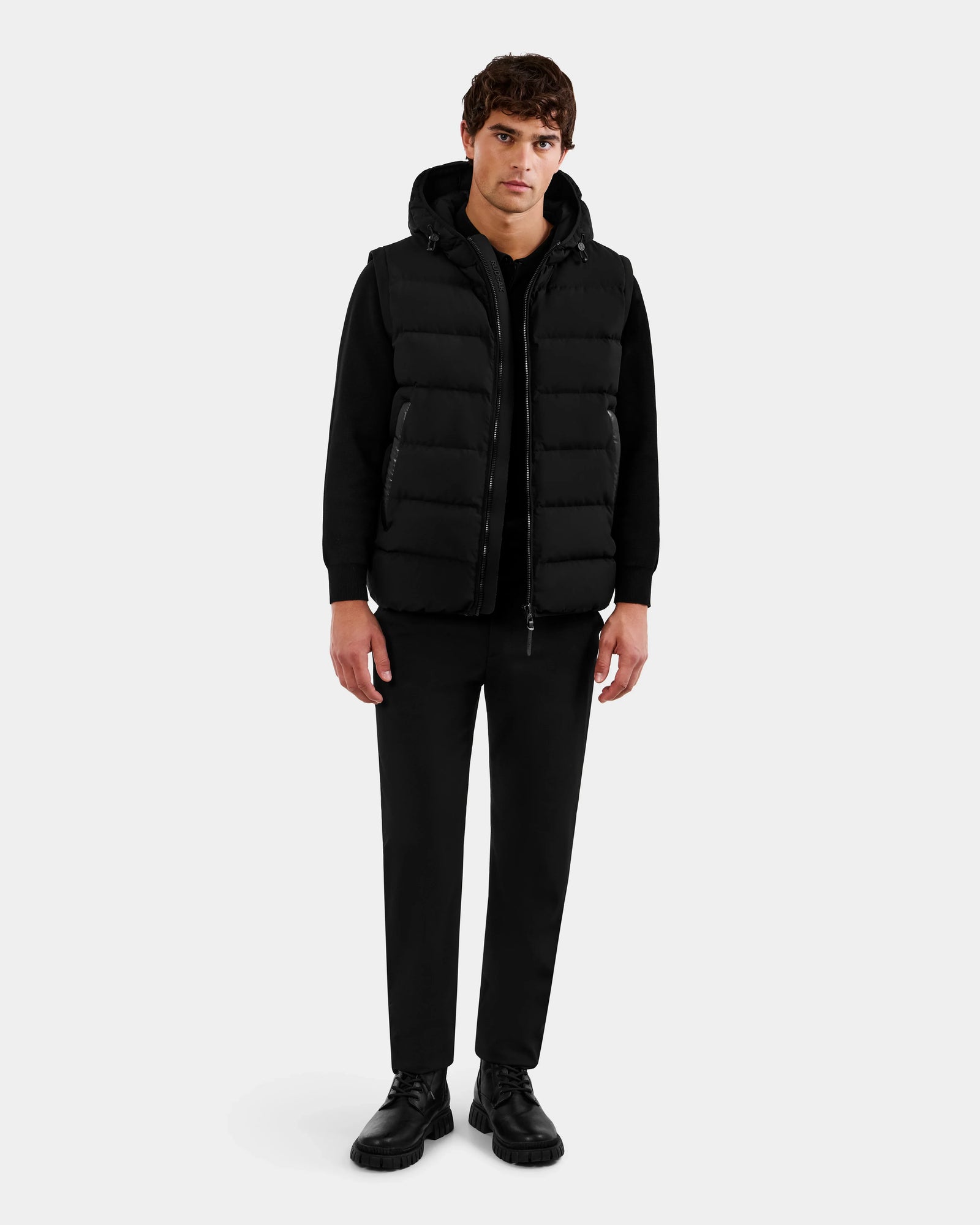 A man faces forward wearing the Rudsak DEMIAN M-R - M123504 Black 2-in-1 puffer vest over a black hoodie, matched with black pants and shoes, set against a plain light gray background.