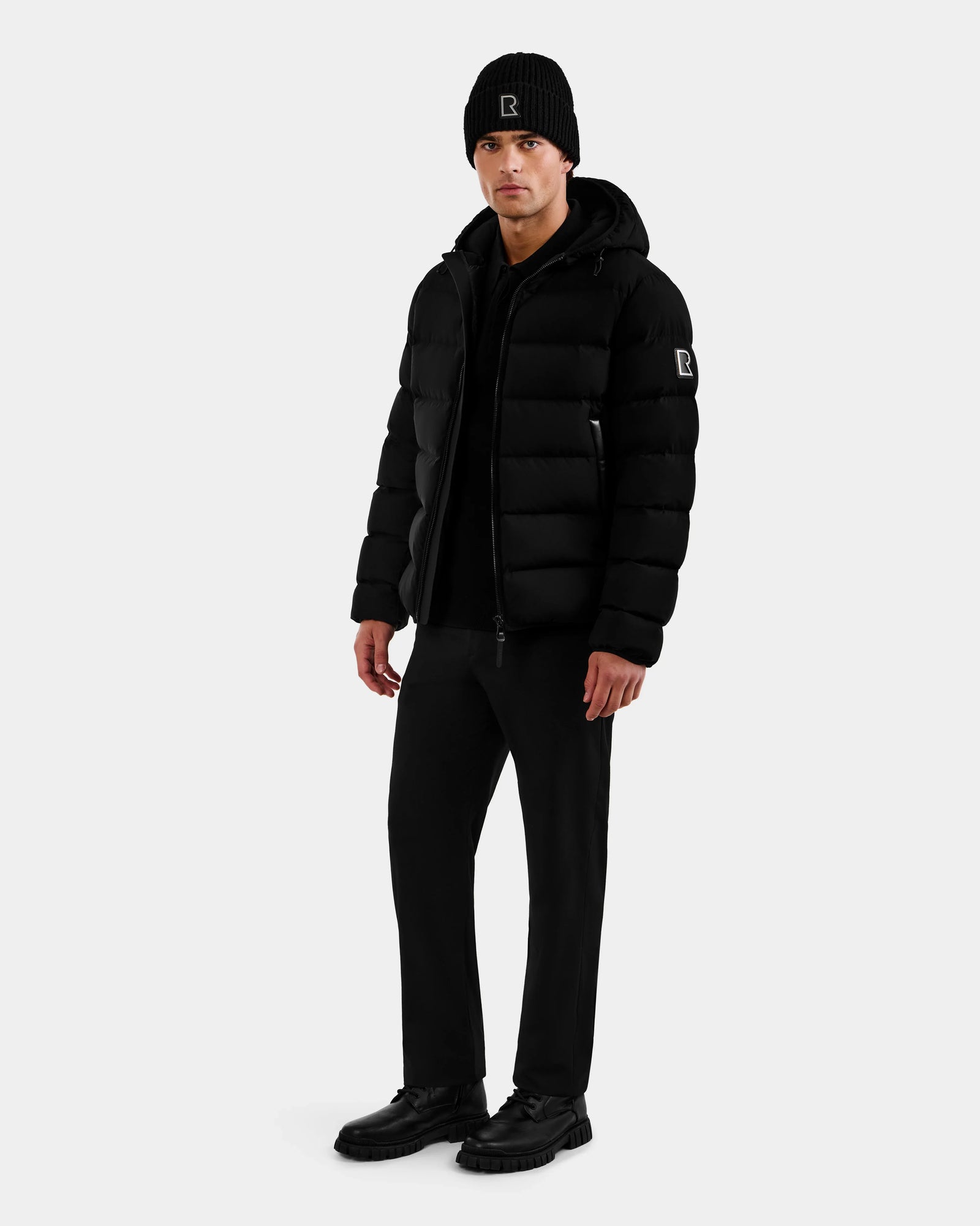 A man in Rudsak’s DEMIAN M - M123507 Black down puffer stands slightly turned to the side, pairing the jacket with a black beanie, pants, and shoes against a plain light background.