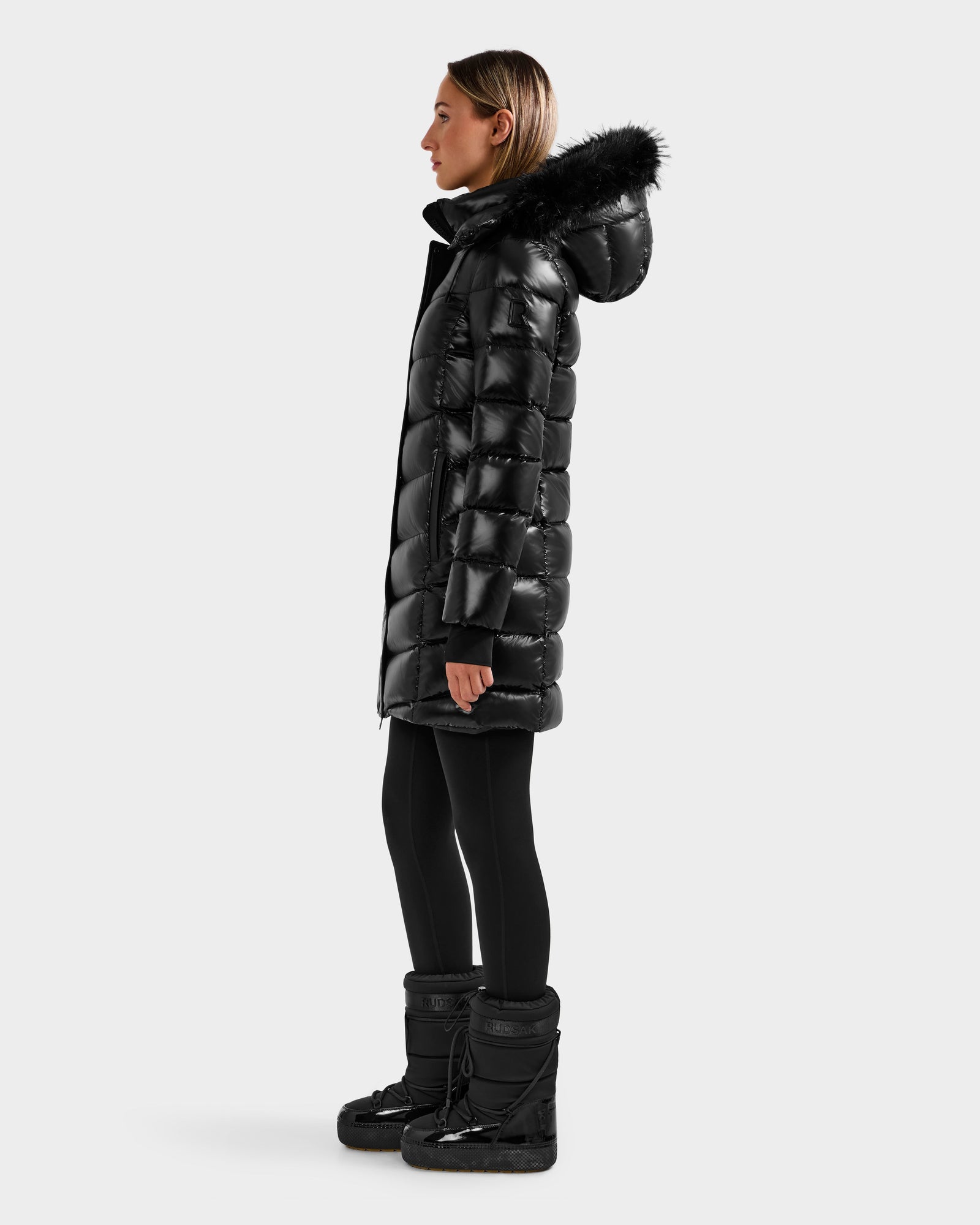 A woman stands in profile wearing the Rudsak ELISA S - L125572 Black quilted puffer jacket with a black fur-lined hood, paired with black leggings and winter boots, against a plain light gray background.