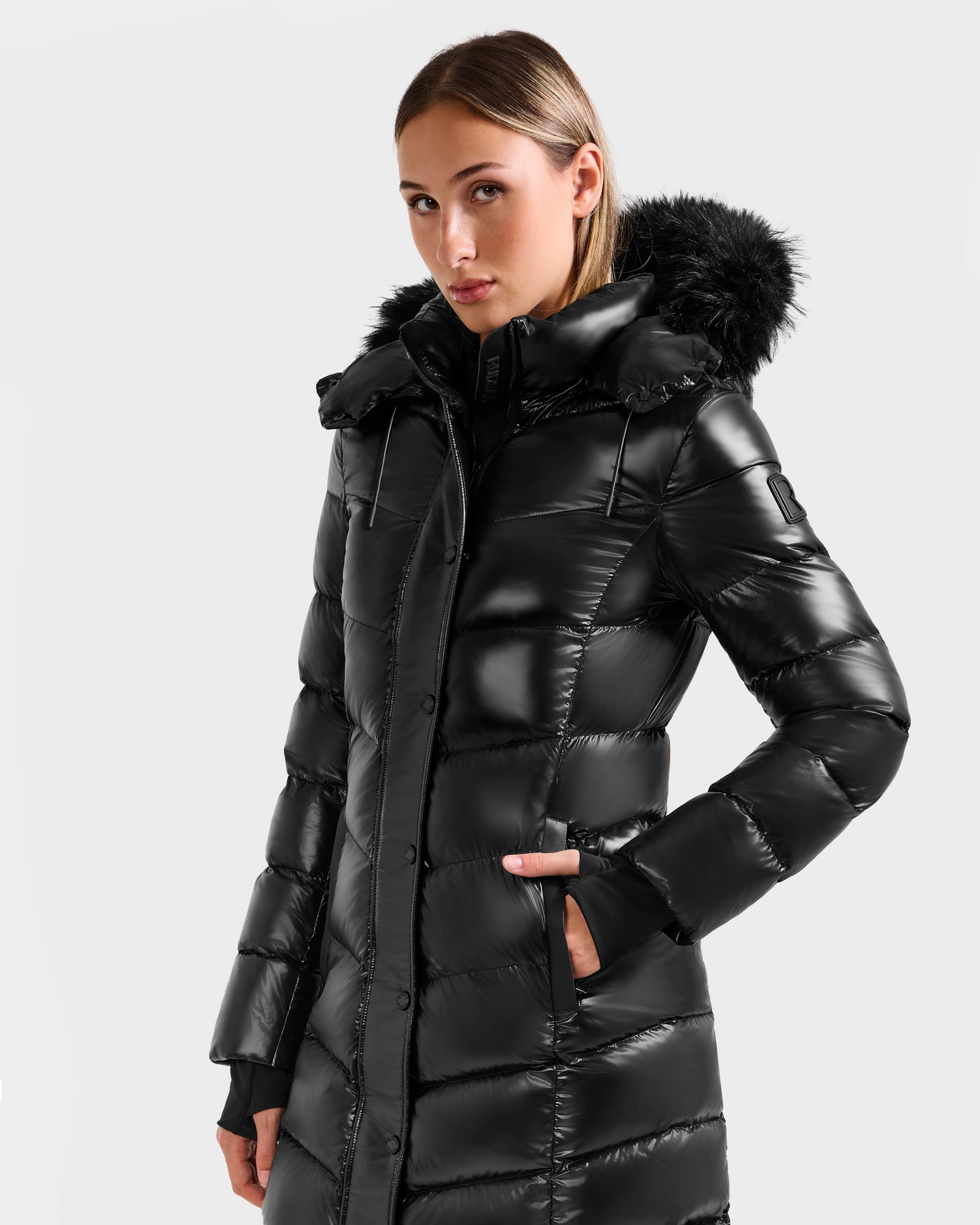 A woman wears the Rudsak ELISA S - L125572 Black down puffer with a fur-lined hood, standing confidently with one hand in her pocket against a plain light background.