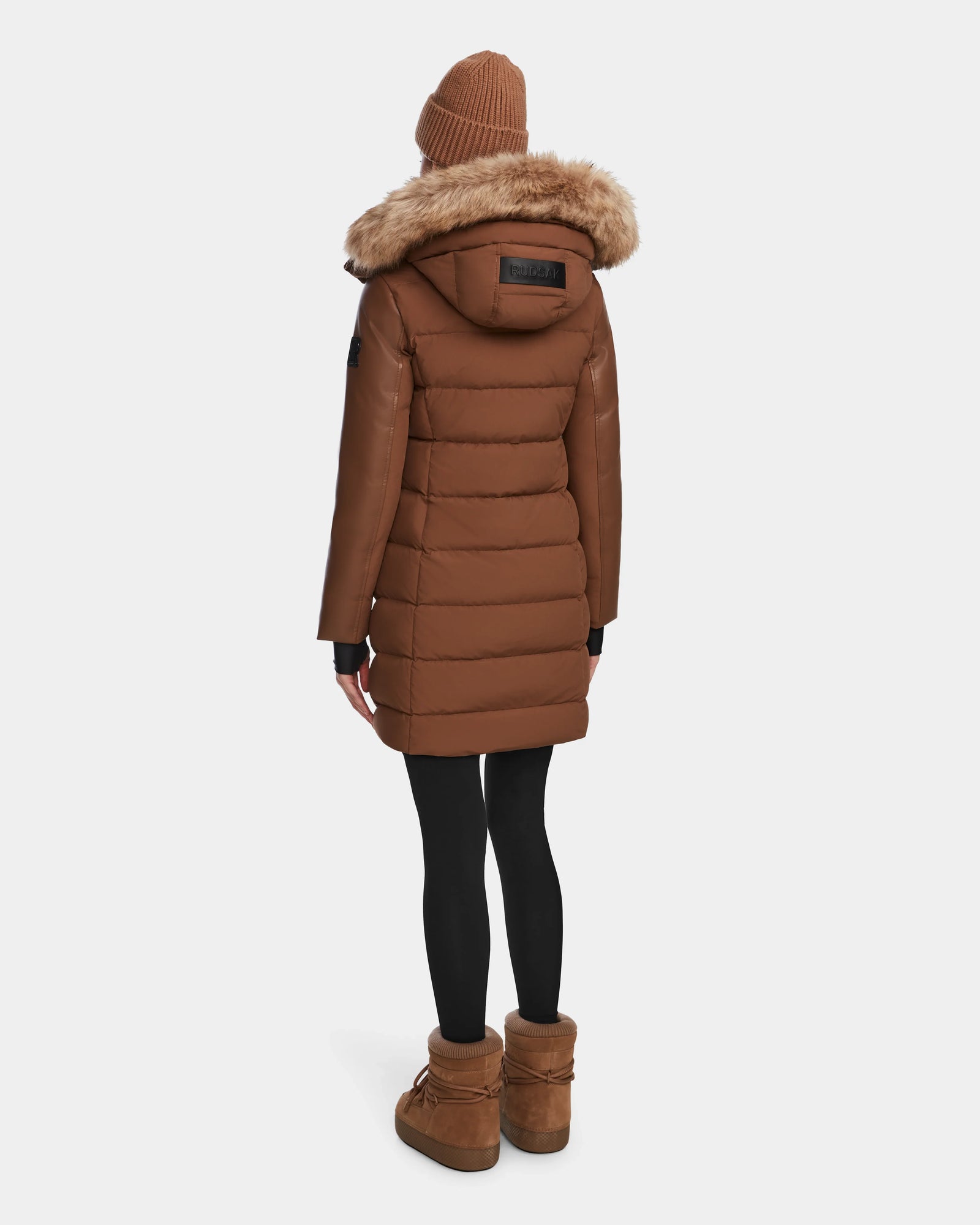 A person wearing the Rudsak ELSA - L123535 Taupe womens quilted down puffer coat with a fur-lined hood faces away from the camera, paired with black leggings, brown boots, and a brown knit beanie against a plain background.
