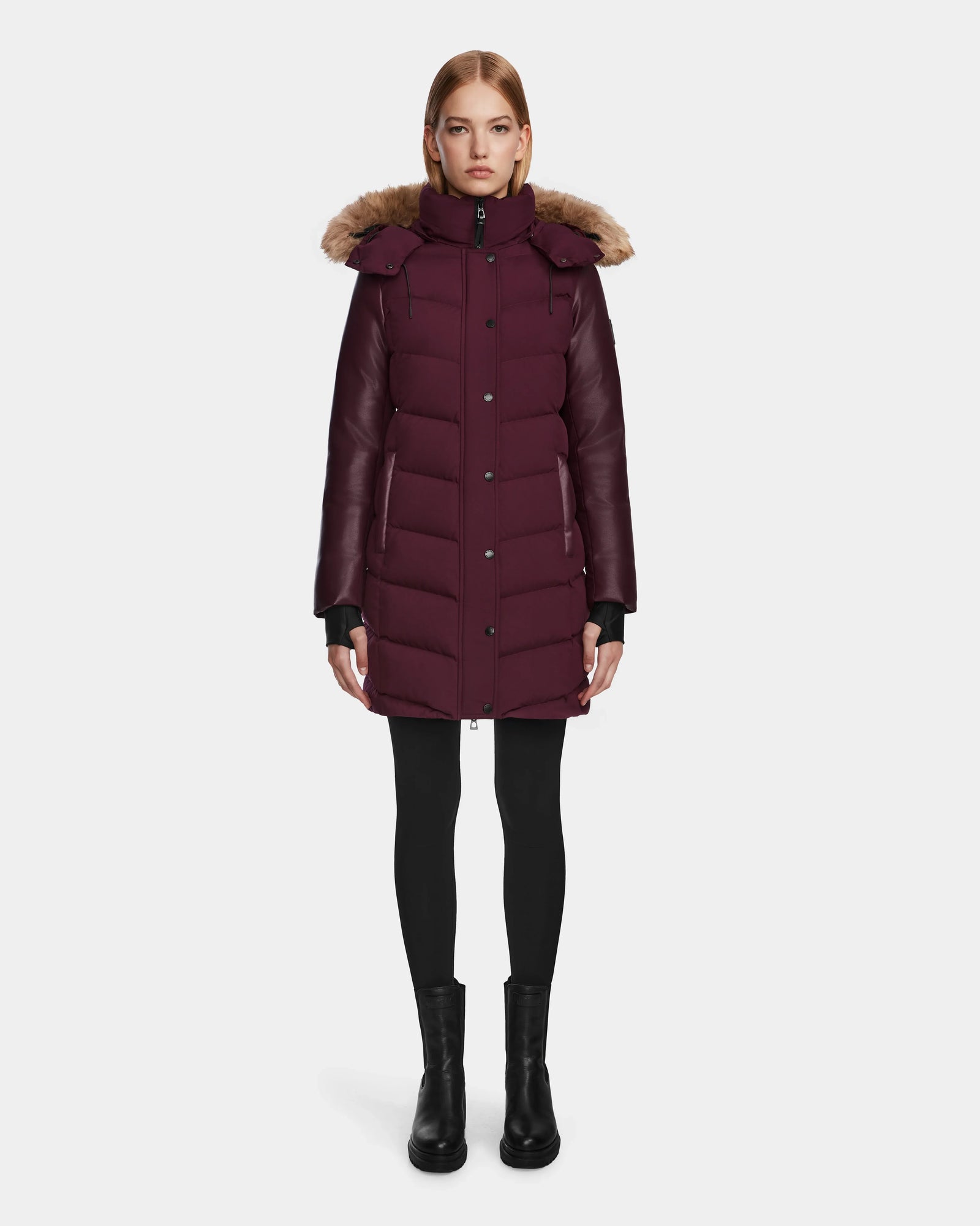 A woman faces forward wearing the Rudsak ELSA - L123535 Wine quilted down puffer with a faux fur-trimmed hood, paired with black leggings and boots, set against a plain light gray background.
